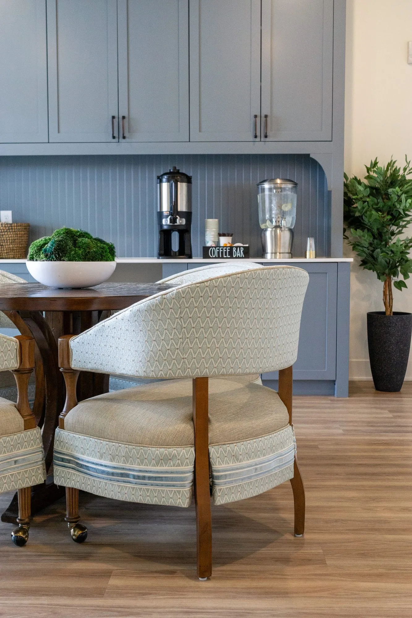 Senior living interior detail: pale blue barrel chair at a walnut table before the grey-blue coffee station and a potted tree