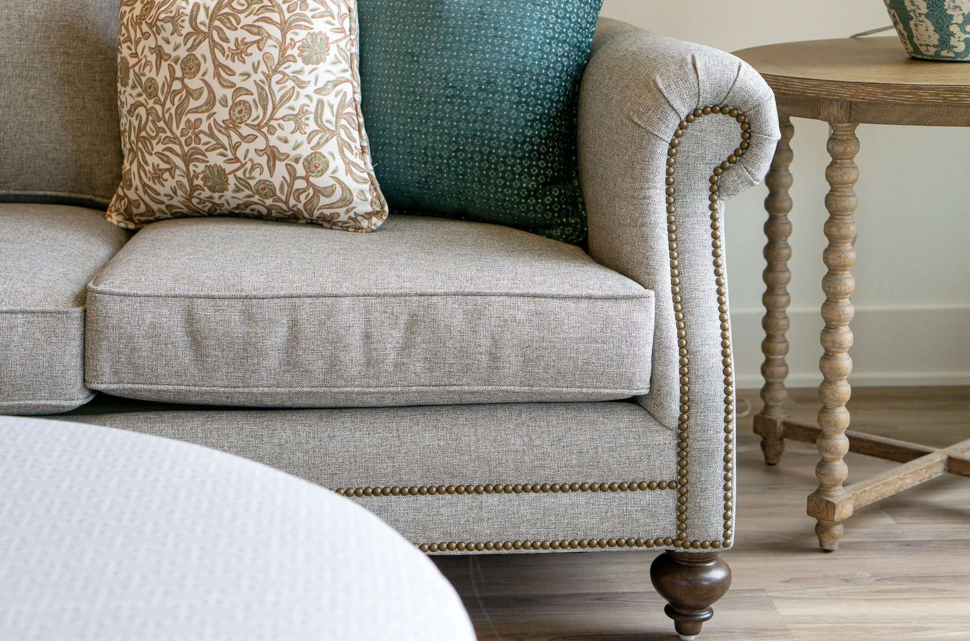 Senior living interior detail: grey sofa's rolled arm with nailhead trim and turned foot beside a spindle table