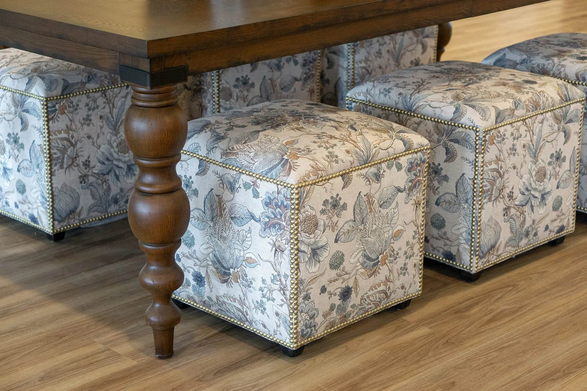 Senior living interior detail: floral cube ottomans with nailhead trim beside a turned walnut table leg, from the side