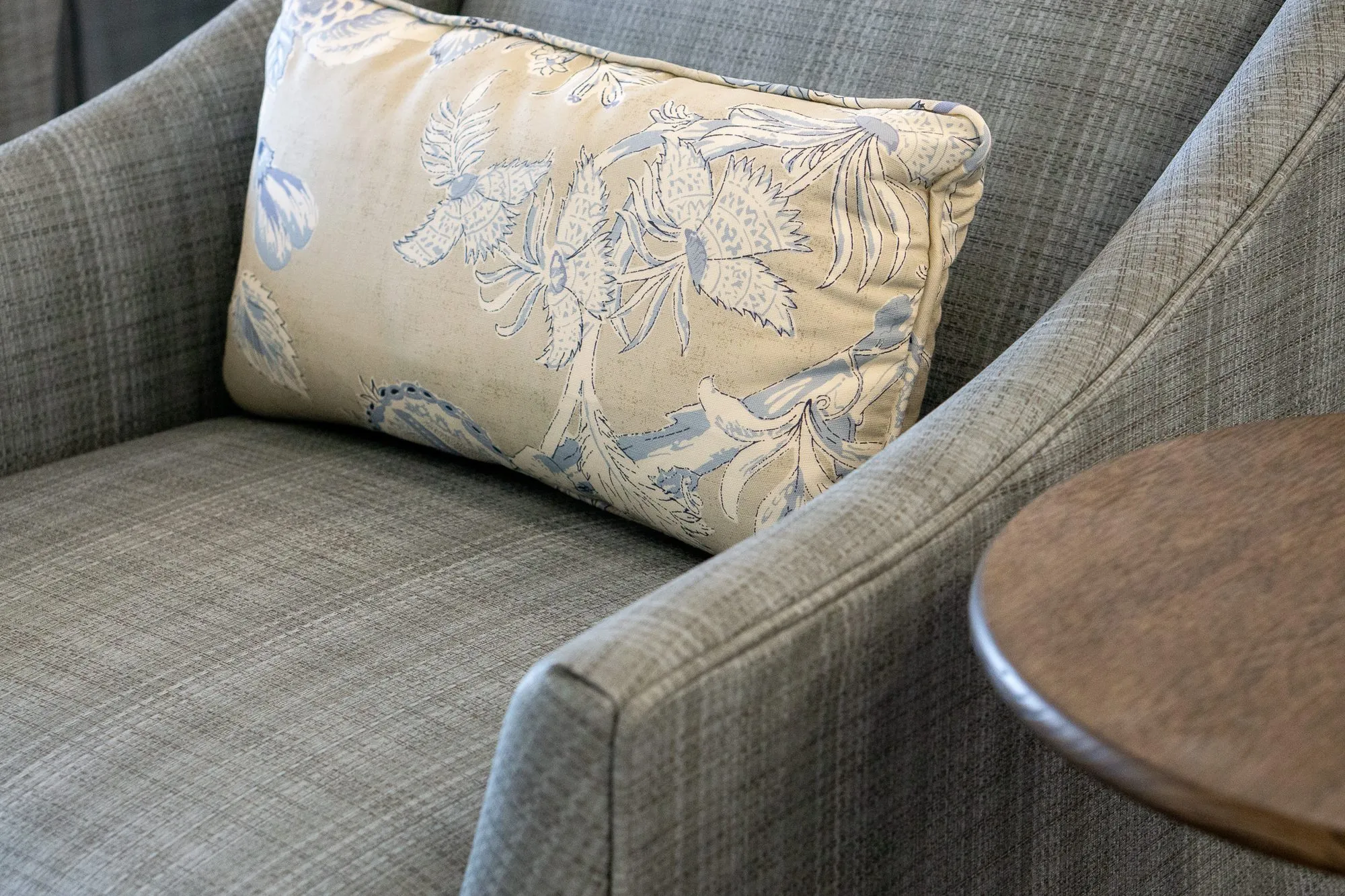 Senior living interior detail: floral lumbar pillow on a grey plaid armchair beside a round drink table