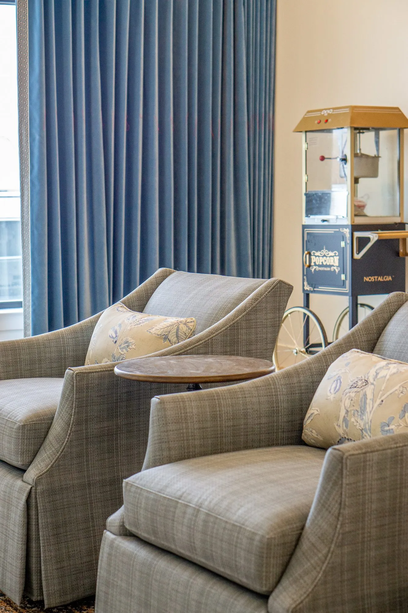 Senior living interior detail: grey plaid armchairs with floral pillows beside blue curtains and a popcorn cart