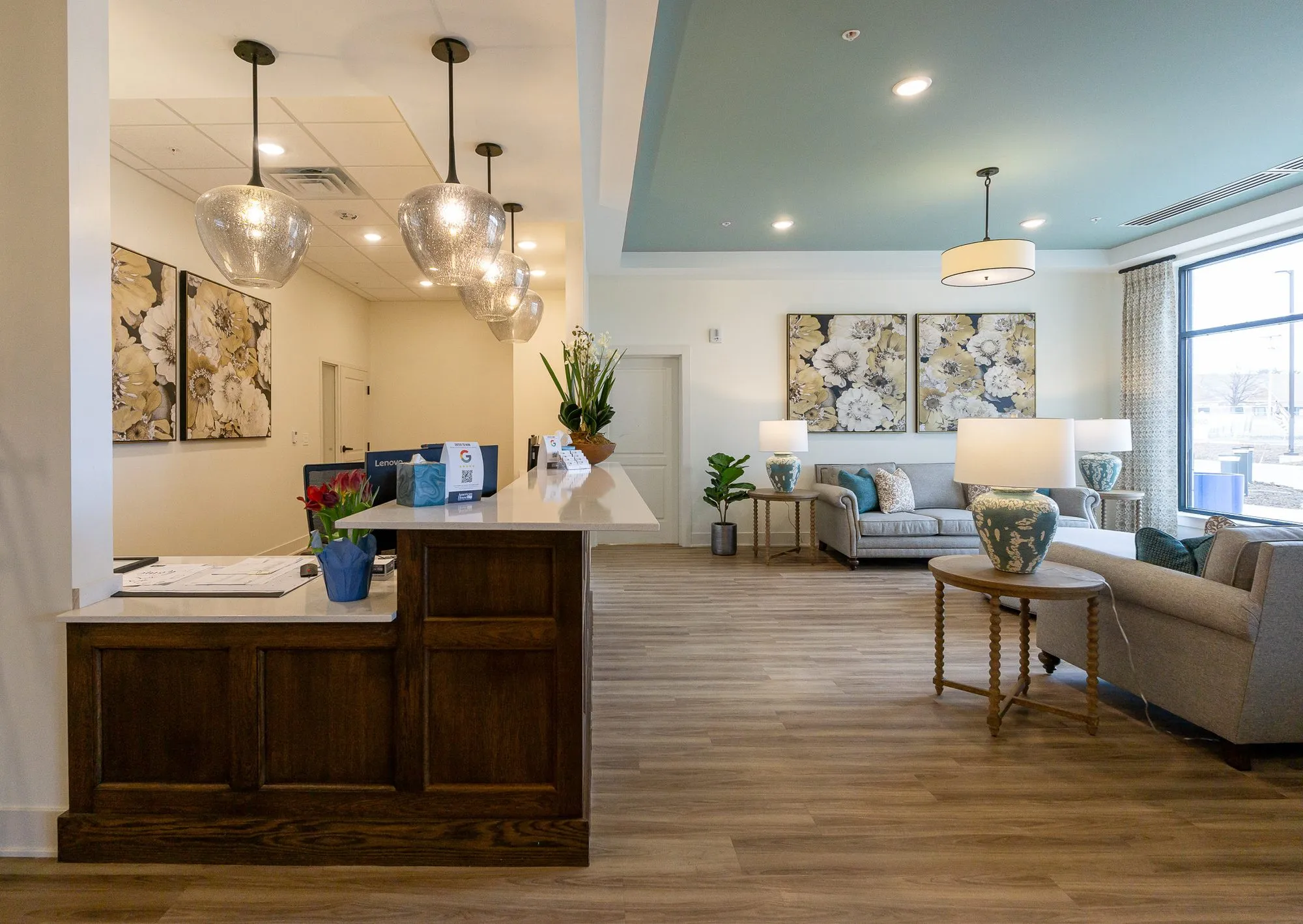 Senior living concierge desk under smoked glass pendants, the lounge with grey sofas and floral canvases at right