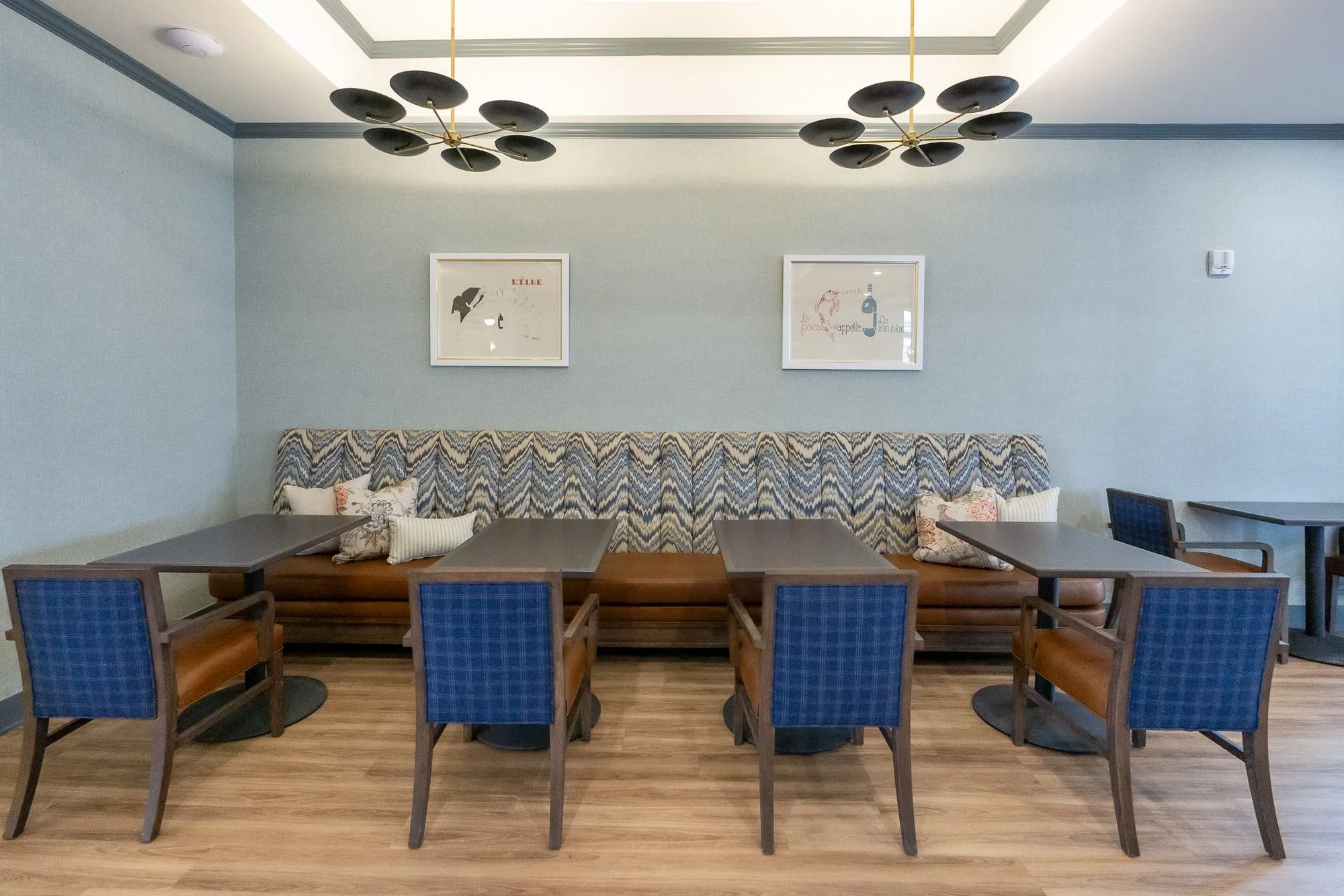 Senior living dining room with blue plaid chairs at tables before the chevron banquette, framed prints and disc chandeliers