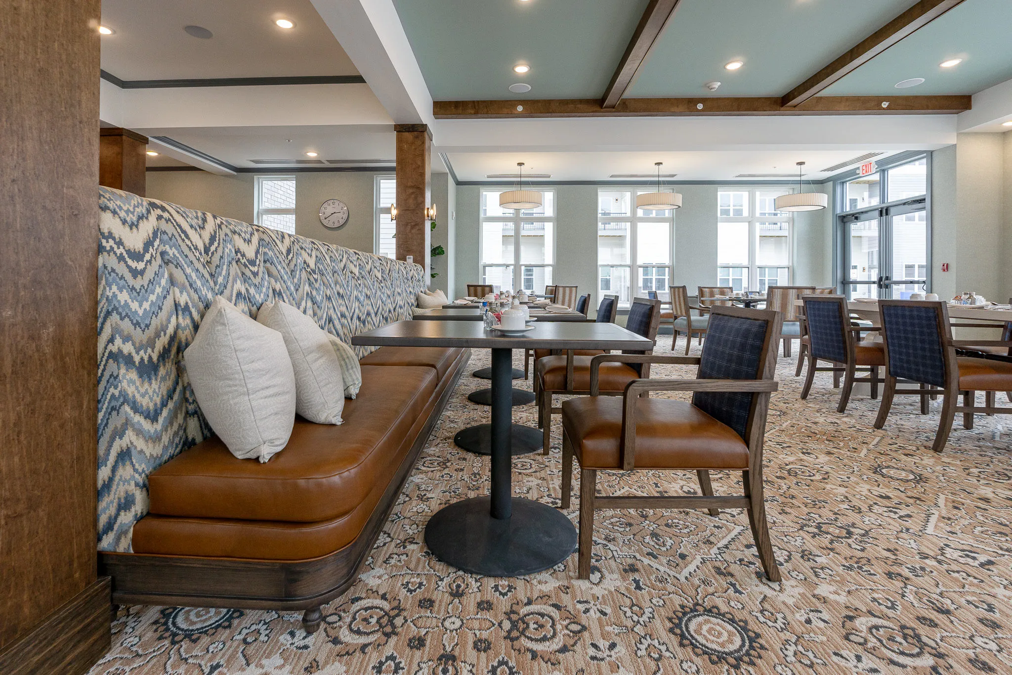 Senior living dining room with a caramel leather banquette with an ikat back, blue plaid chairs at tables and timber columns