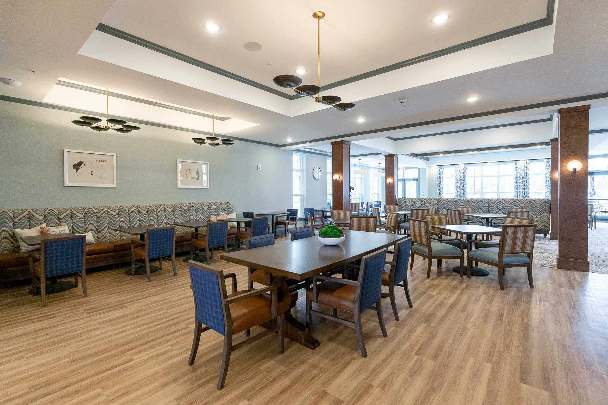 Senior living dining room with a chevron banquette and blue plaid chairs at tables under black disc chandeliers, tray ceiling