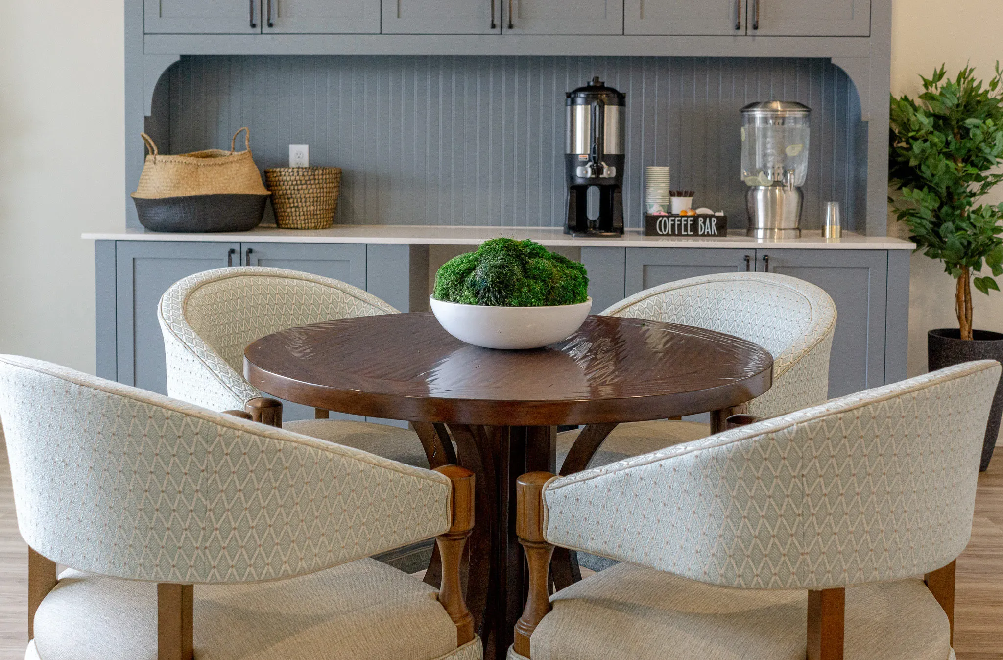Senior living coffee bar with cream barrel chairs at a round table facing the grey-blue servery and urns