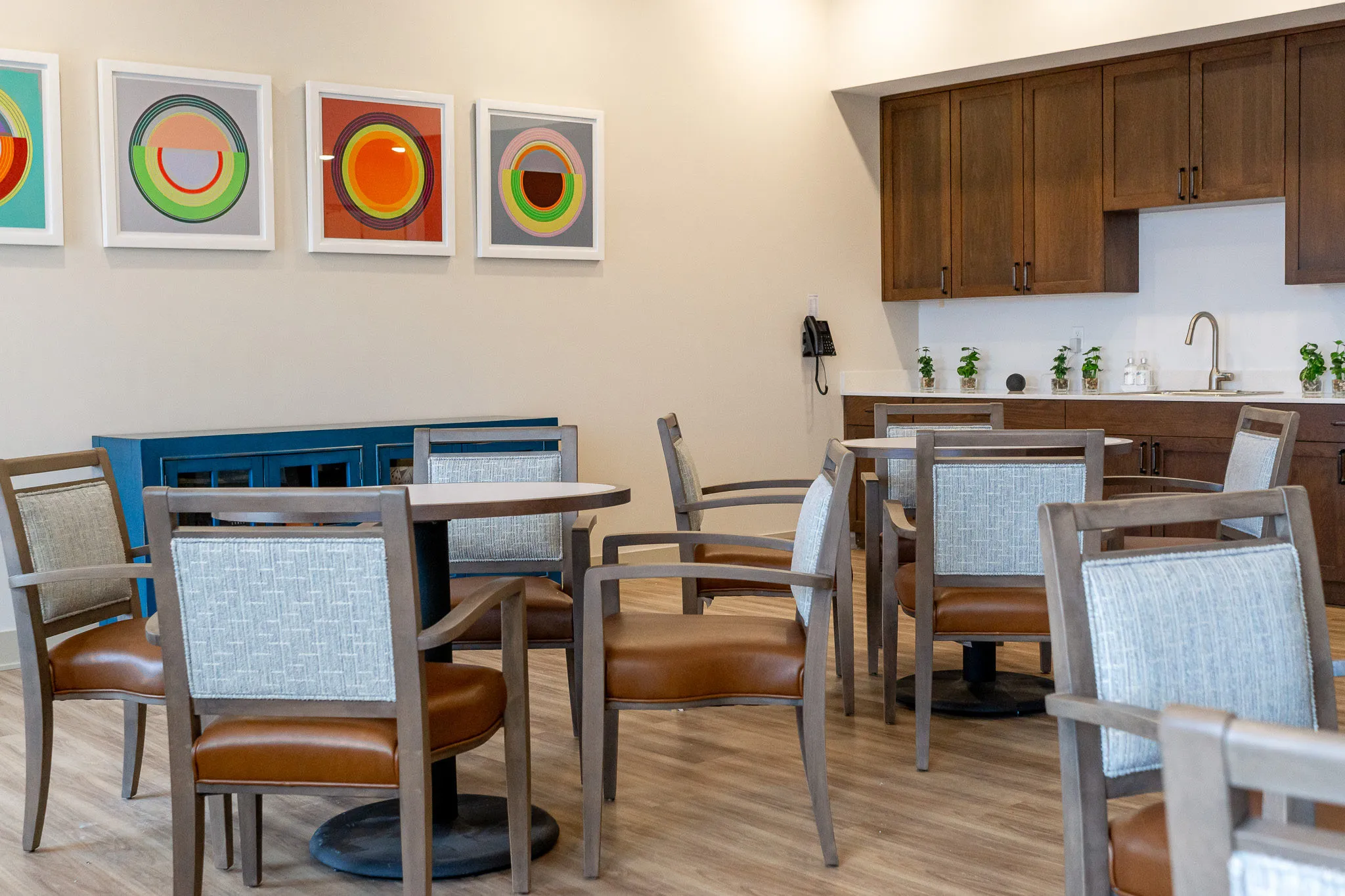 Senior living game room with card tables and leather-seated chairs beside a kitchenette and a row of pop art prints