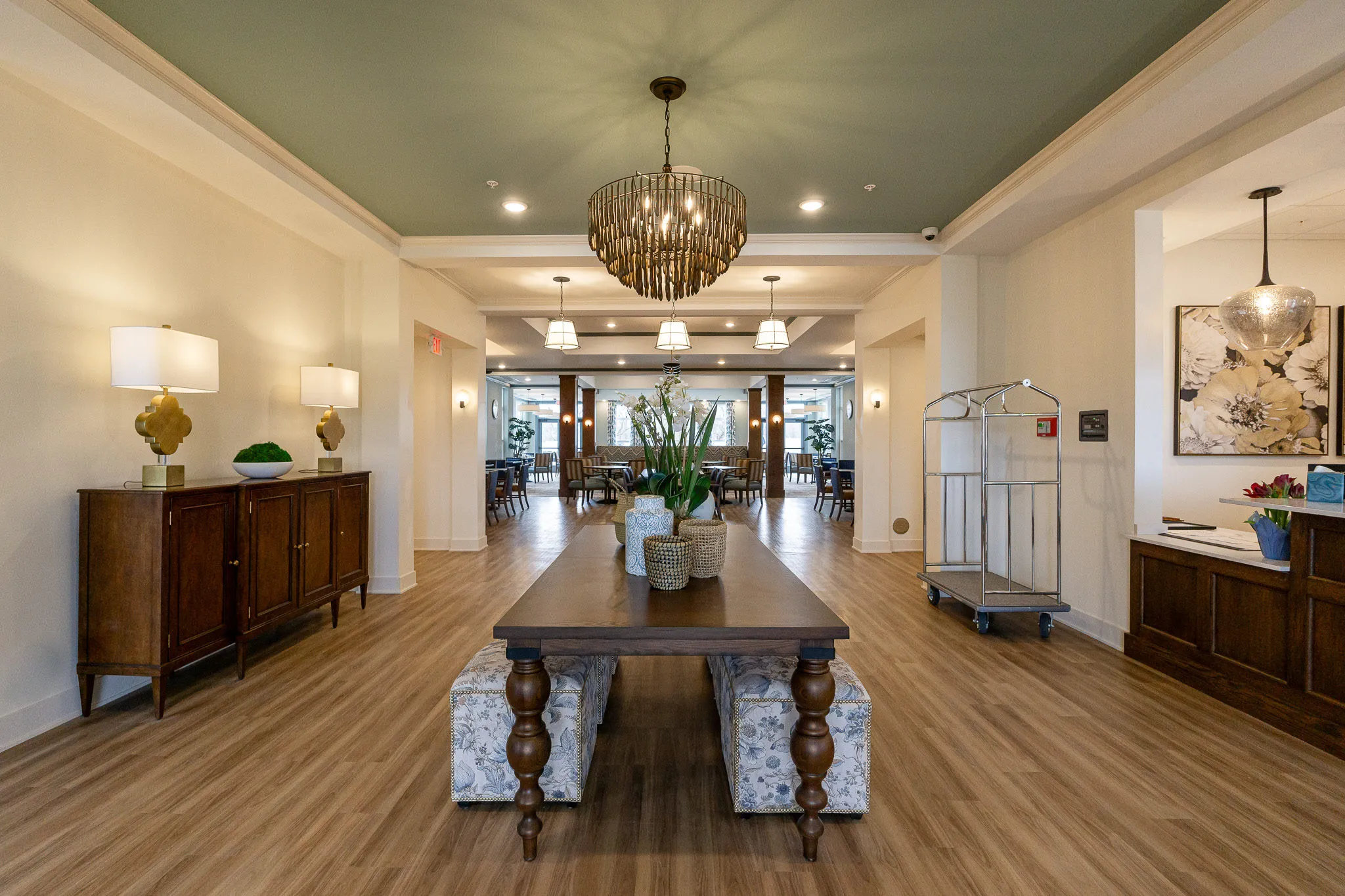 Senior living grand corridor with a console table and floral ottomans below a beaded chandelier, the dining room beyond