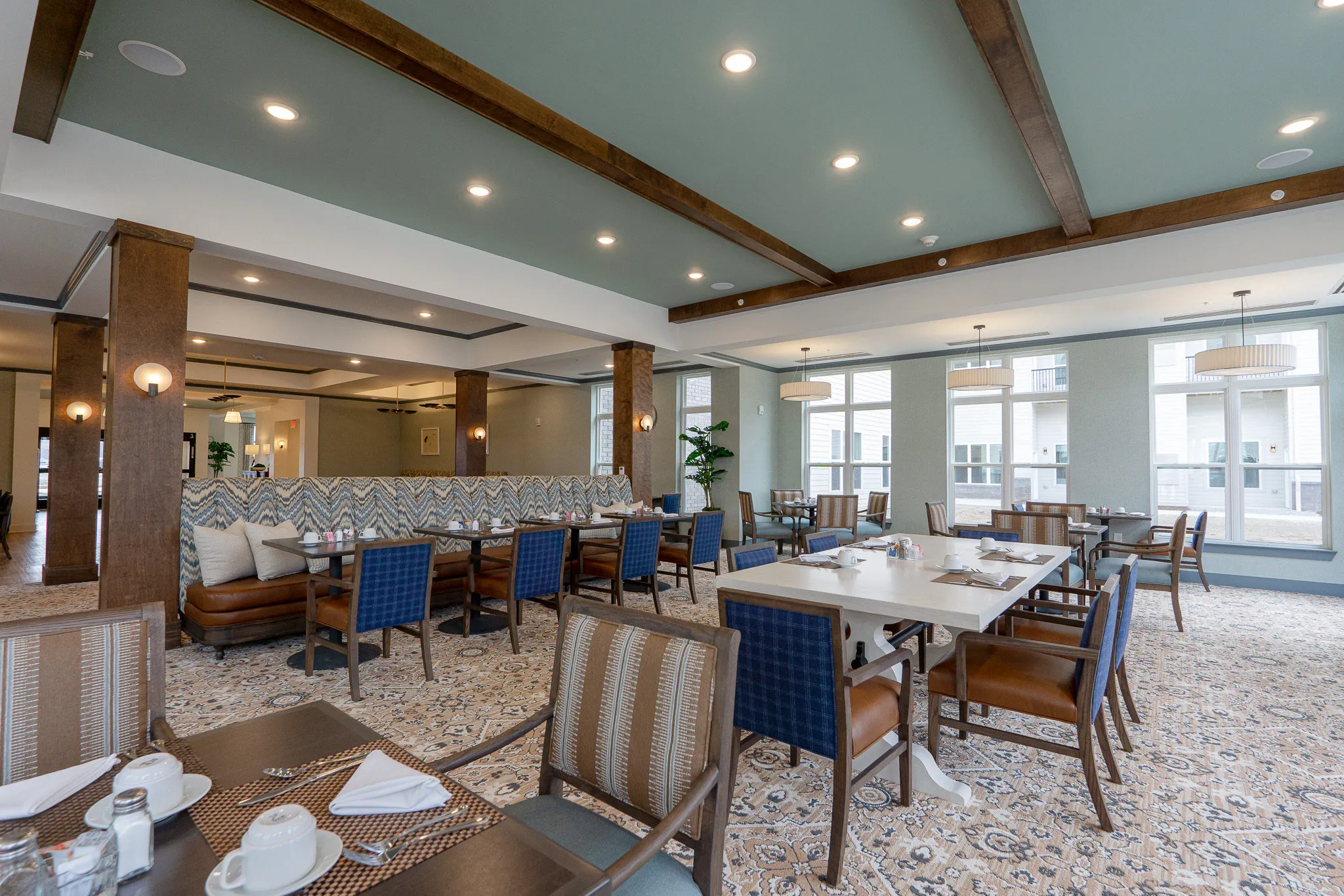 Senior living dining room with blue and striped chairs around a white long table, timber columns and a chevron banquette