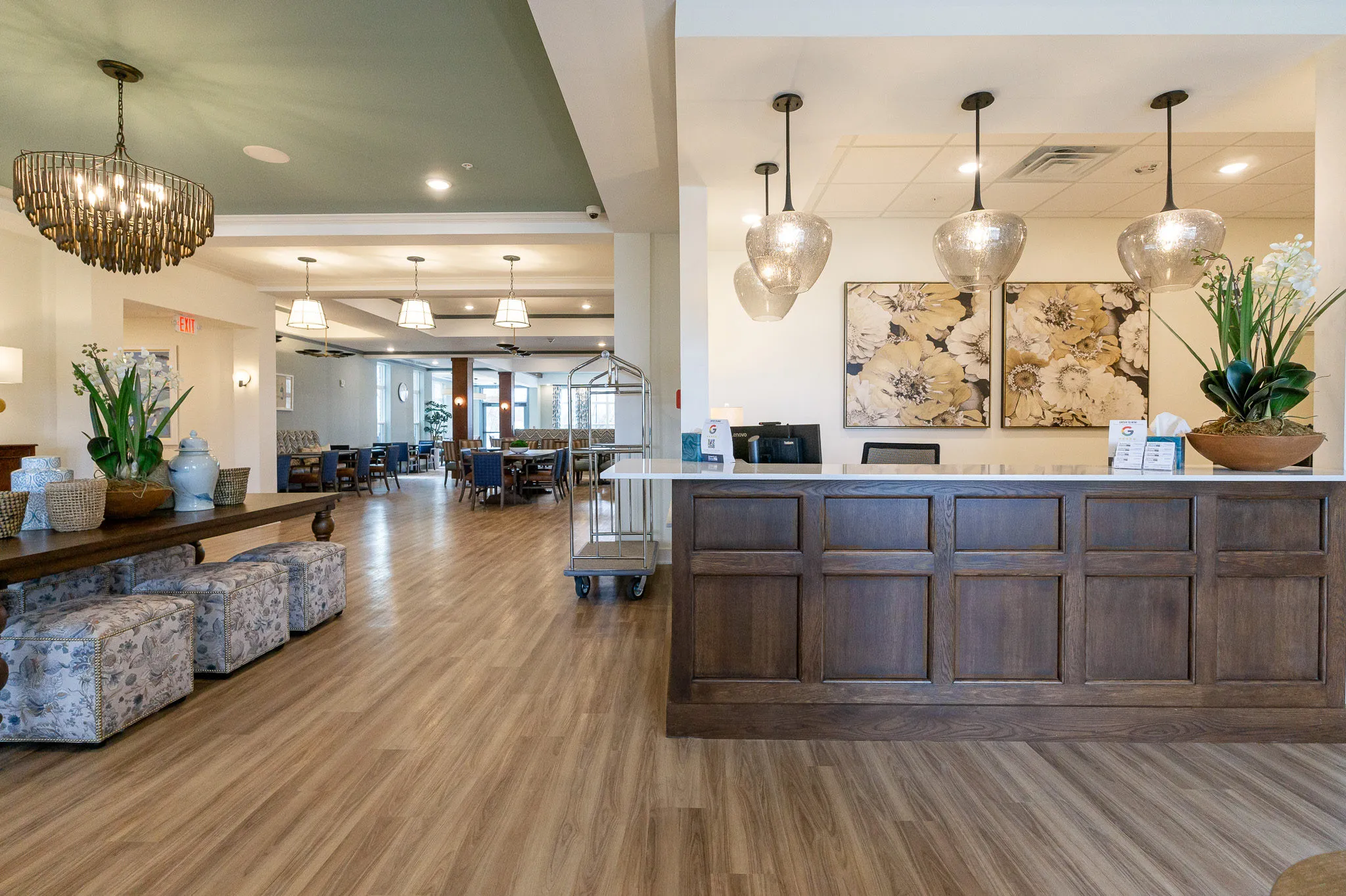 Senior living lobby with a walnut concierge desk below mercury glass pendants beside floral canvases