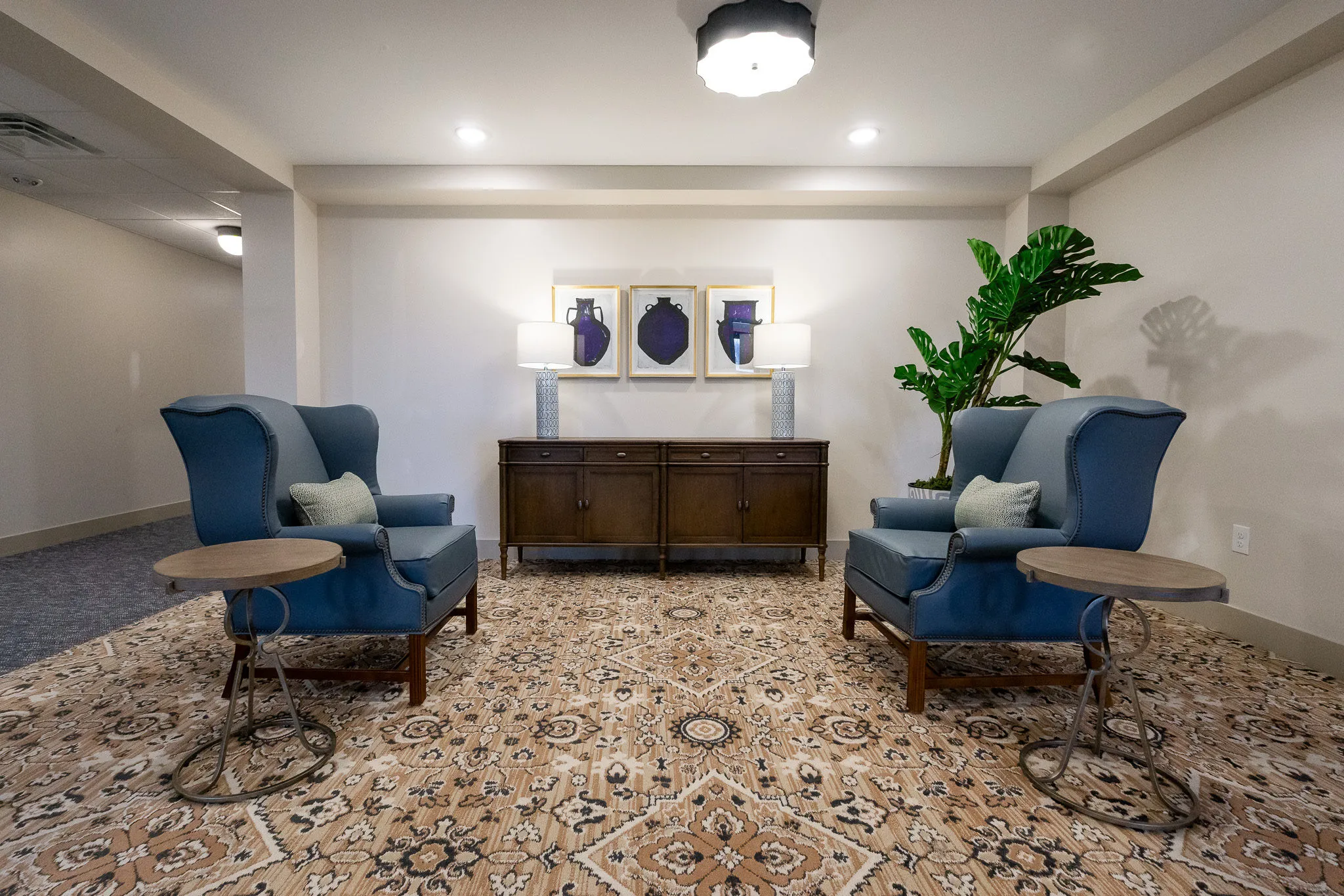 Senior living sitting area with two blue leather wing chairs flanking a walnut console on a patterned carpet