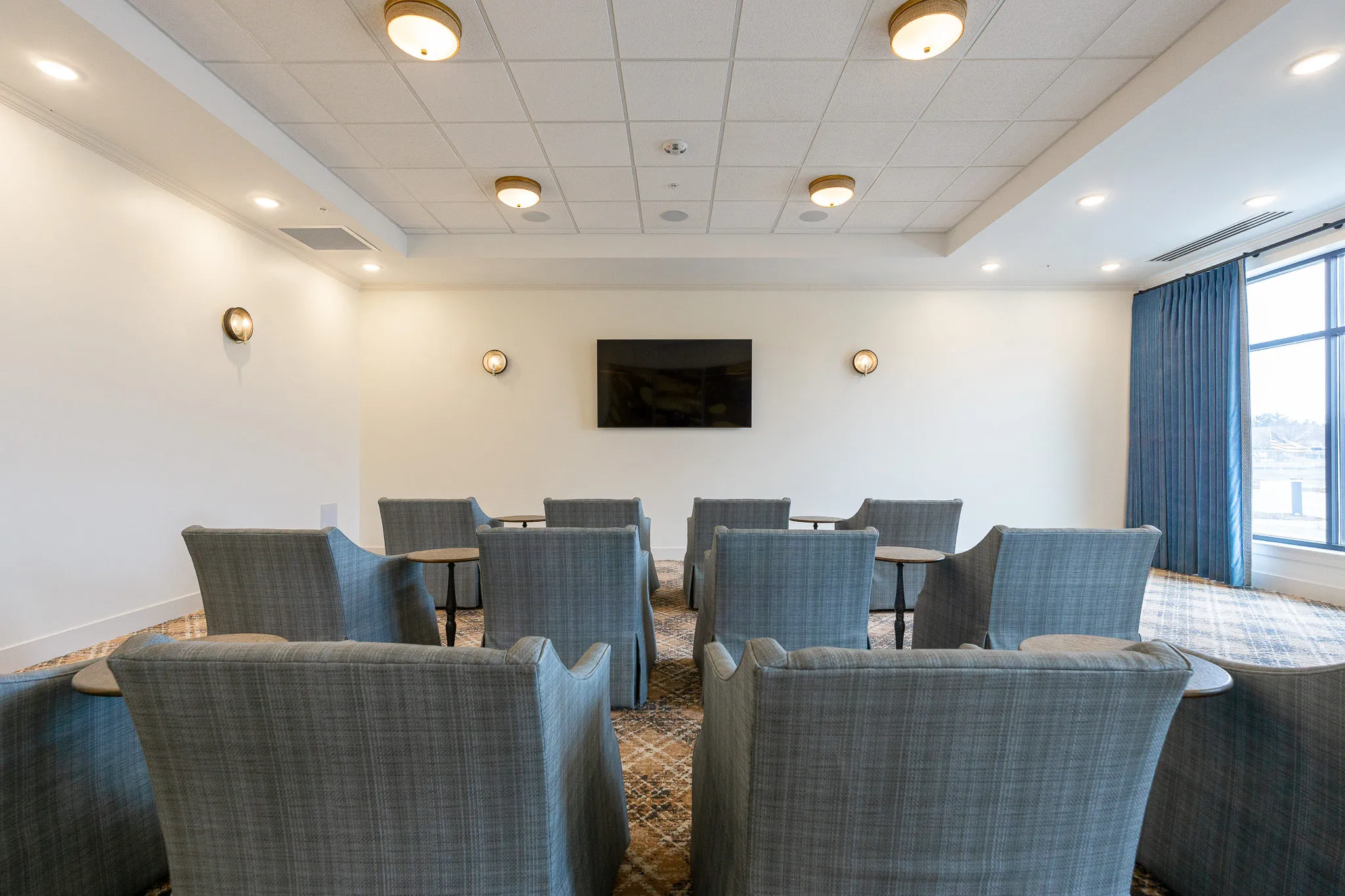 Senior living theater with rows of grey skirted armchairs in pairs around drink tables facing the wall-mounted screen