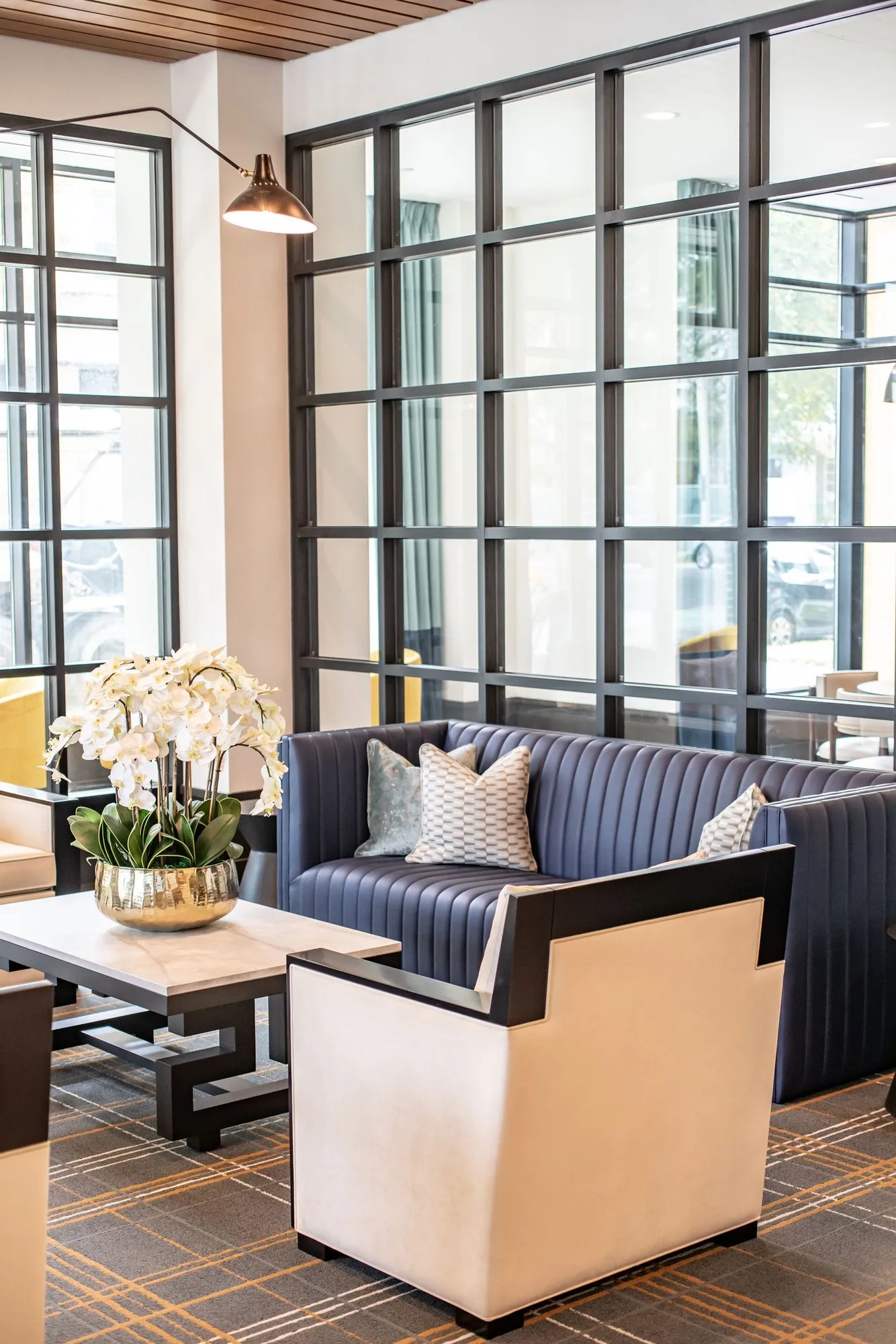 Senior living lounge with a navy channel tufted sofa and white armchair before black steel glazing, orchids on a marble table