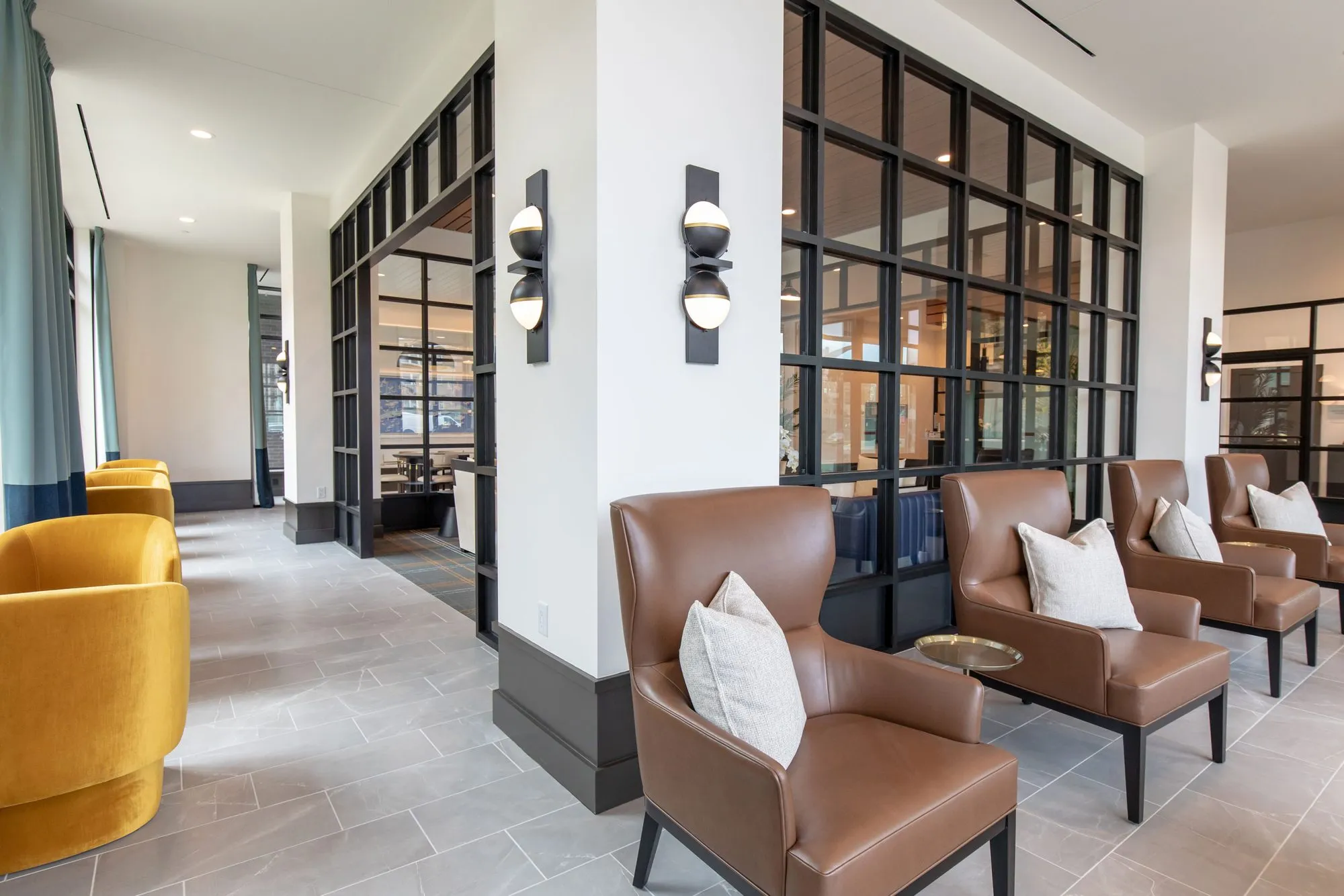 Senior living corridor with cognac leather armchairs and mustard velvet chairs beside black steel glass partitions