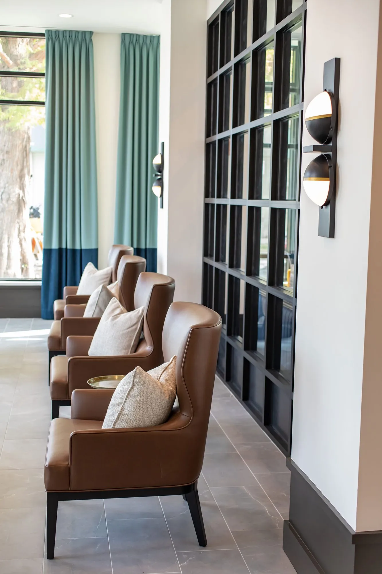 Senior living sitting area with a row of cognac leather armchairs along a black grid partition, curtains beyond