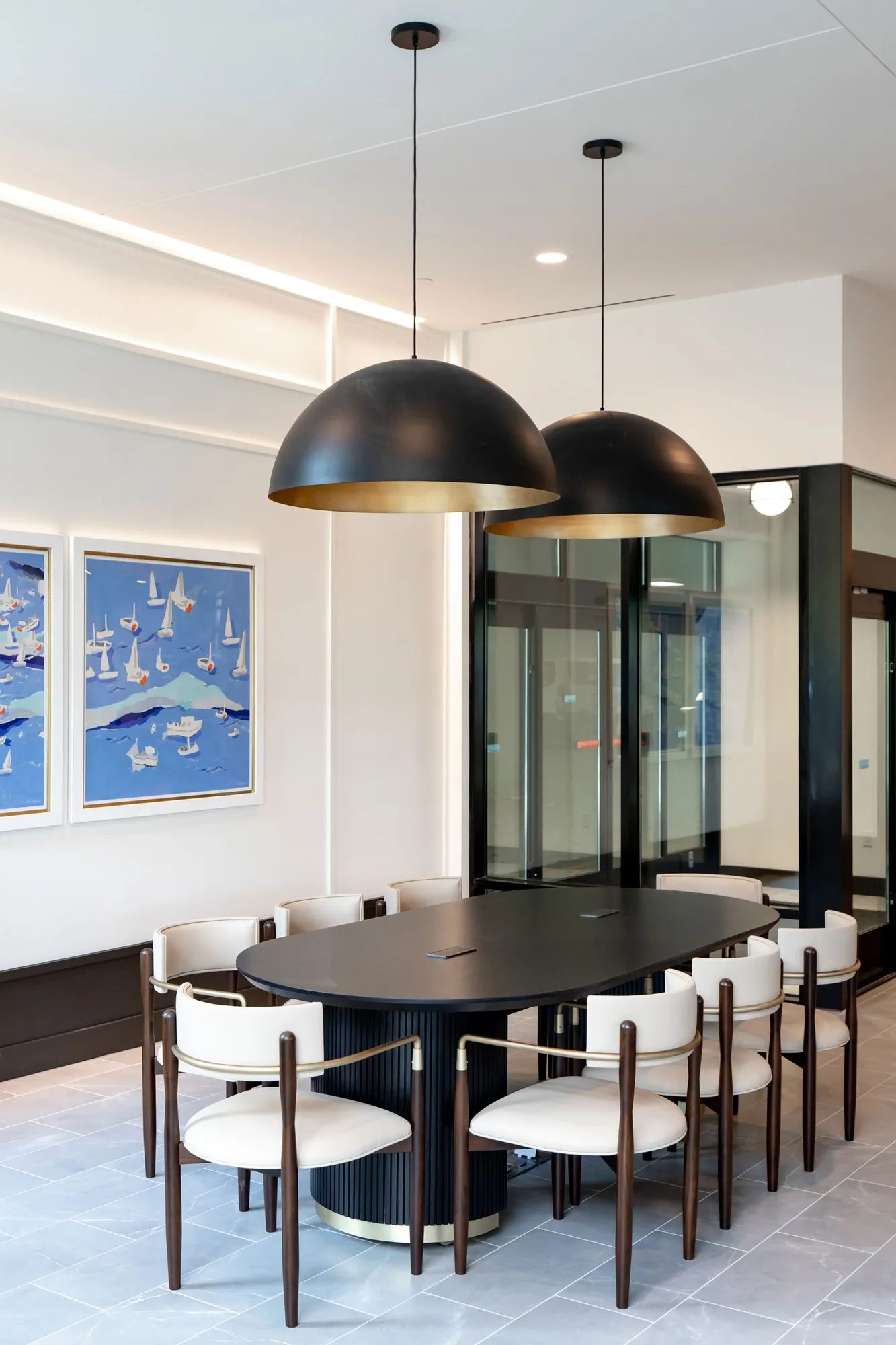 Senior living conference room with a black oval table and cream chairs under two dome pendants, sailboat paintings