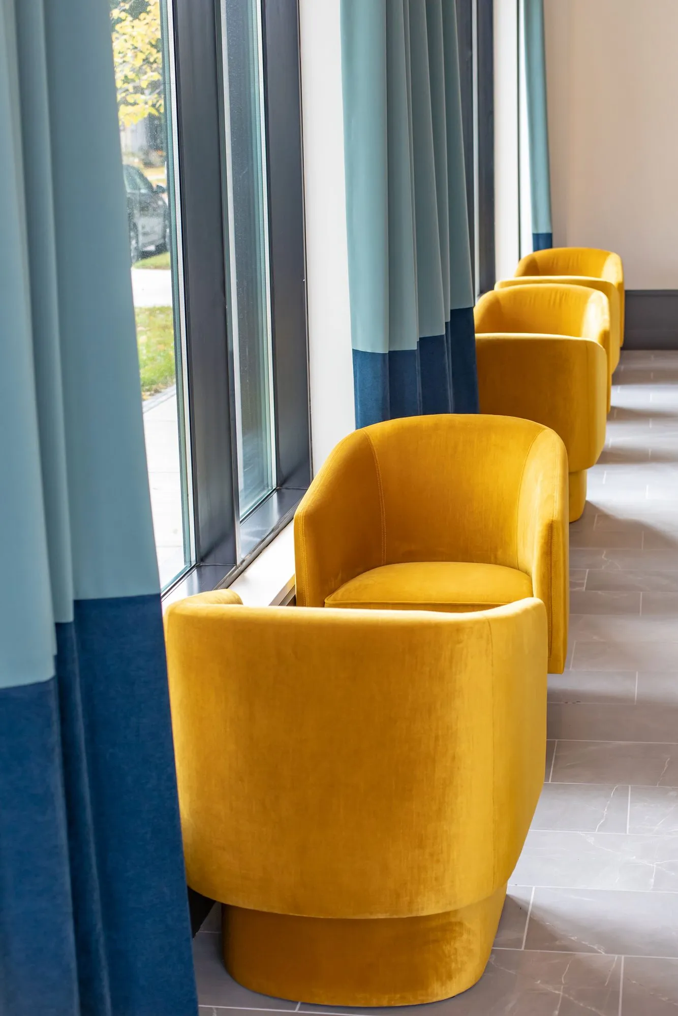 Senior living sitting area with a row of mustard velvet swivel chairs along tall windows hung with teal and navy curtains