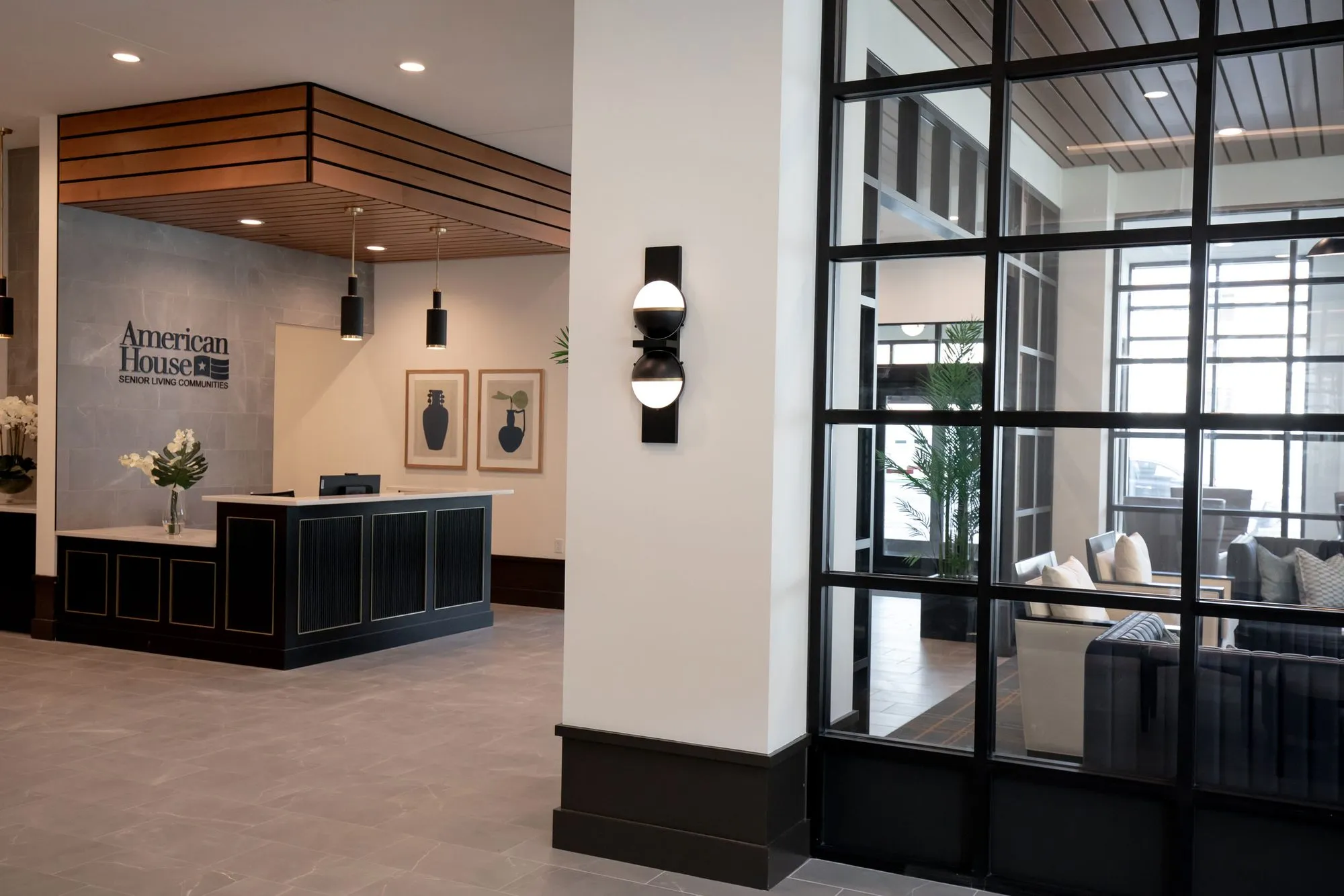 Senior living reception desk under a wood slat canopy beside a globe sconce, the lounge seen through black steel glazing