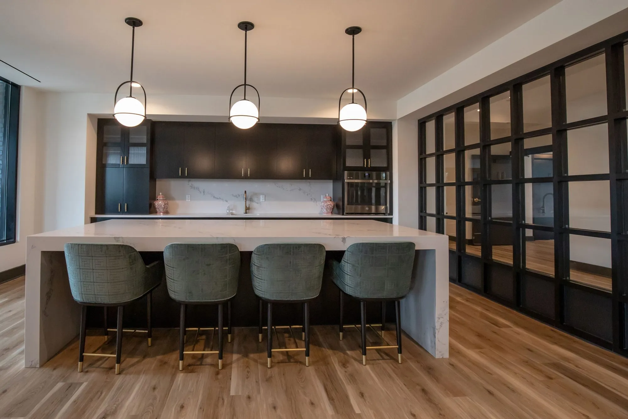 Senior living kitchen island with four green velvet stools under globe pendants, dark cabinetry and a marble backsplash