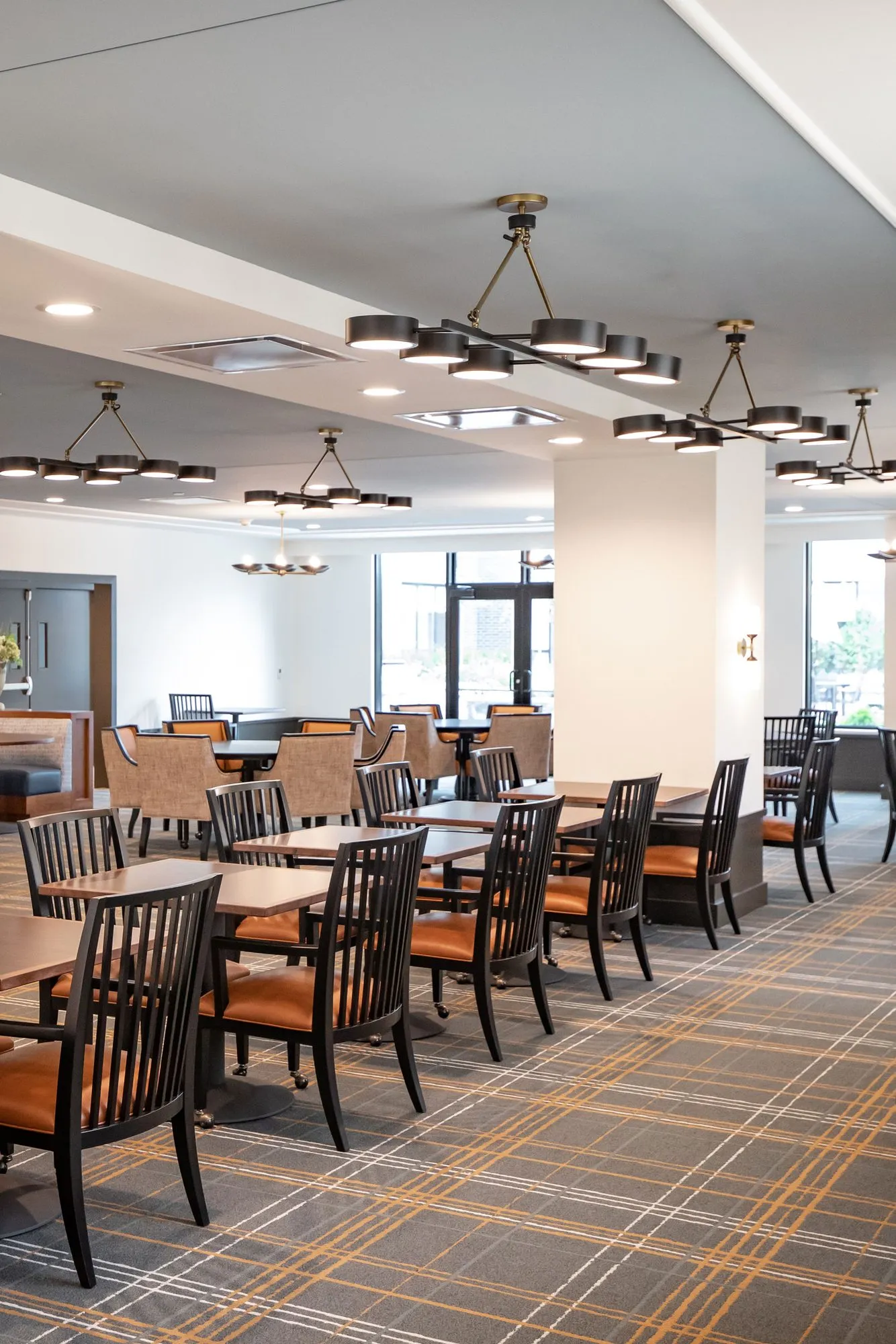 Senior living dining room with black spindle chairs with orange seats under ring chandeliers on plaid carpet