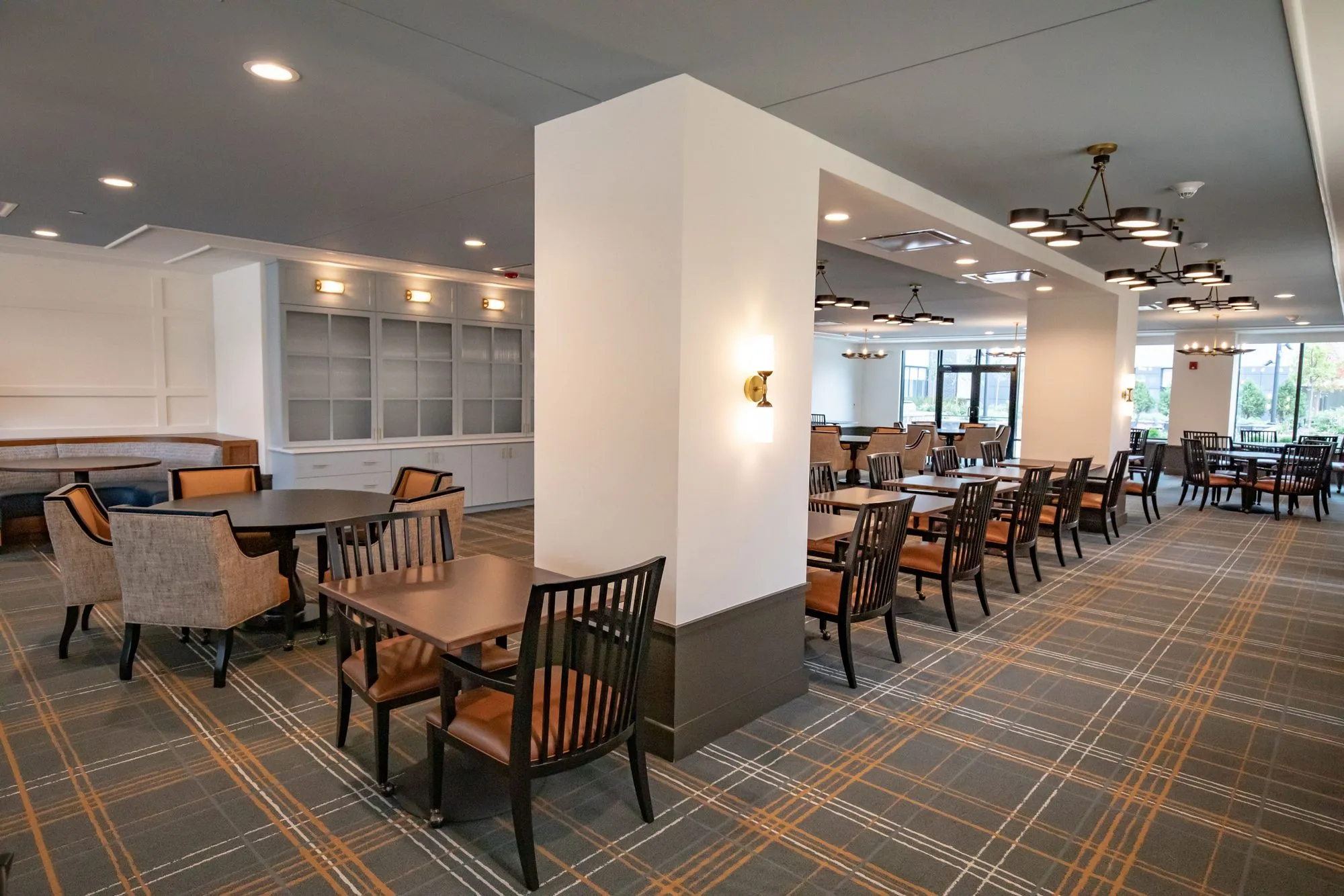 Senior living dining room around a white column with a sconce, woven chairs at a round table and spindle chairs