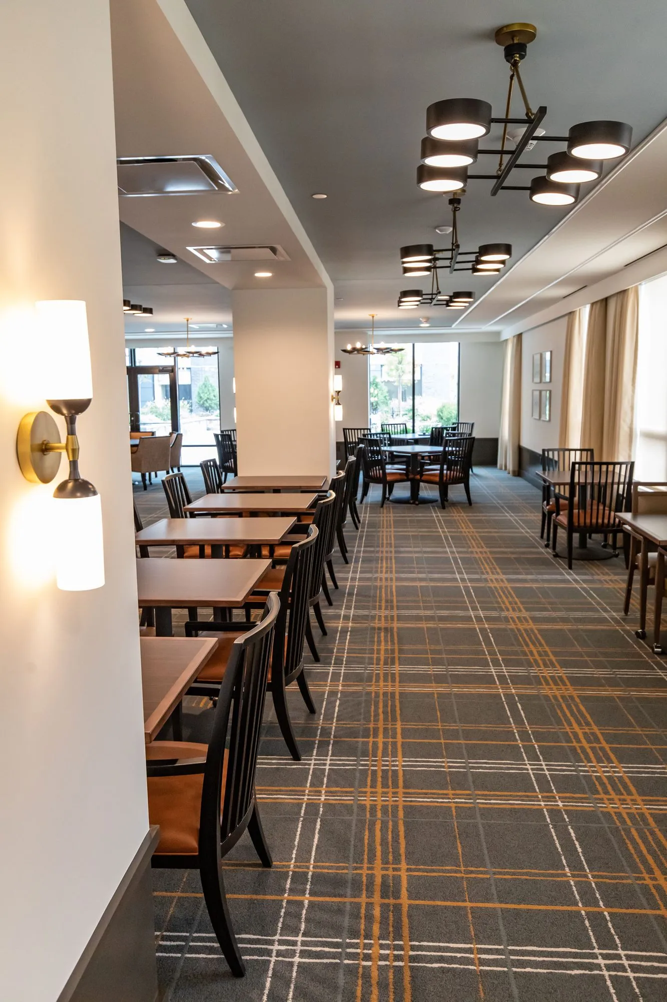 Senior living dining room in a long view, spindle chairs at tables on plaid carpet under ring chandeliers