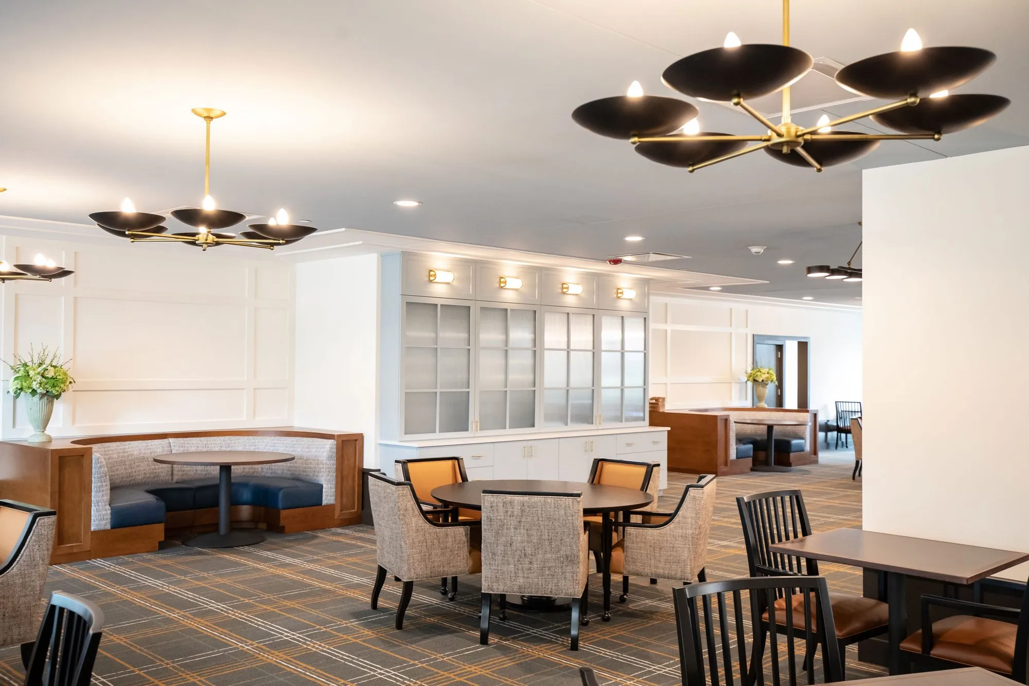 Senior living dining room with a navy curved banquette and upholstered chairs under black and brass saucer chandeliers