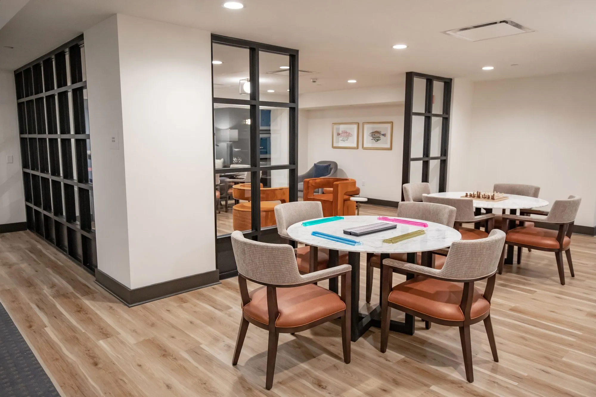 Senior living activity room with tan leather chairs around round tables, one with a chess board, beside black steel openings