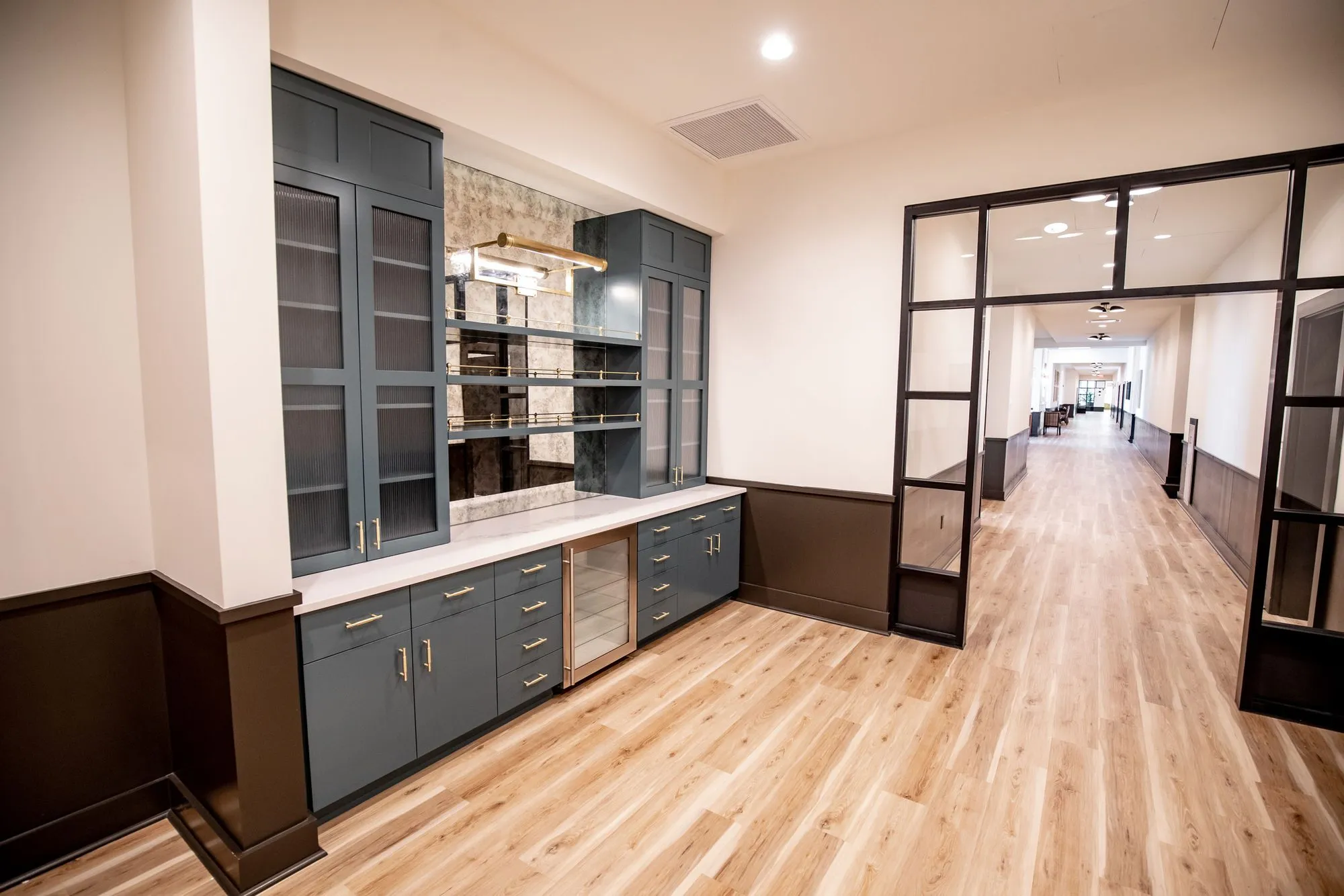 Senior living kitchenette with grey cabinetry, glass front uppers and a wine fridge beside a glazed opening to a corridor