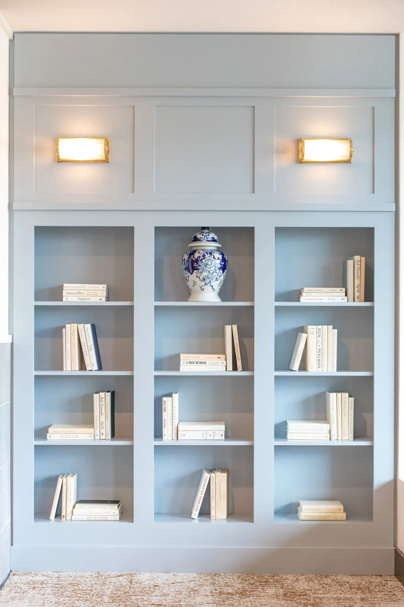Senior living interior detail: pale blue built-in bookcase with white books and a ginger jar below two brass sconces