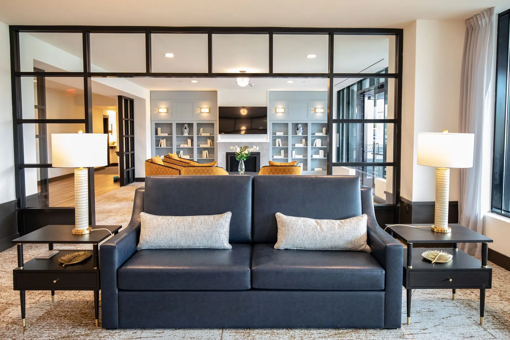 Senior living lounge with a navy leather sofa between white lamps on black tables, a fireplace and bookcases through glazing