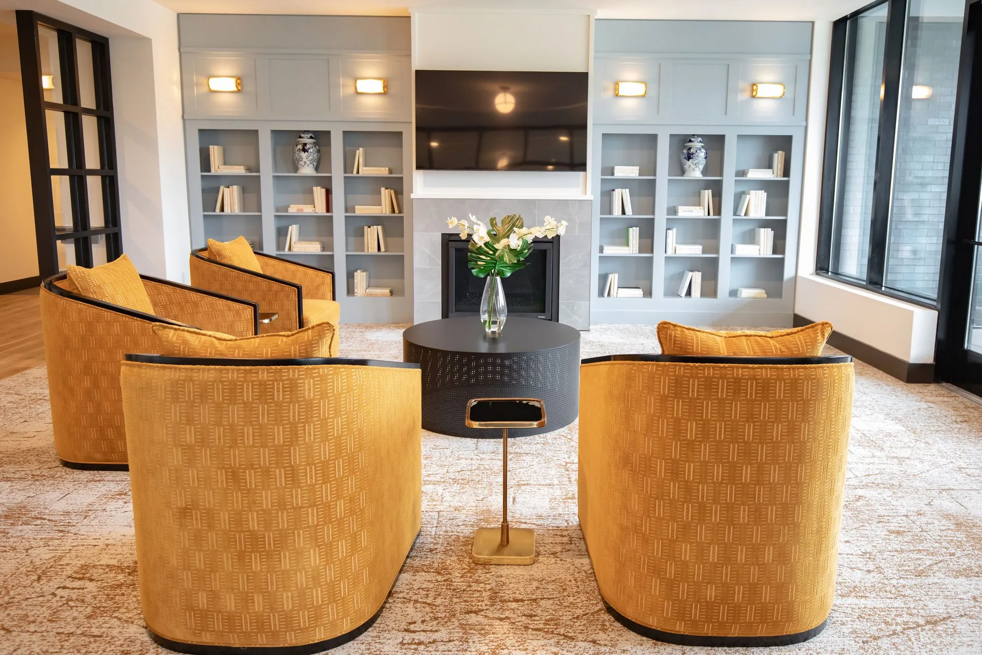 Senior living lounge with mustard swivel chairs around a black drum table facing a fireplace between pale blue bookcases