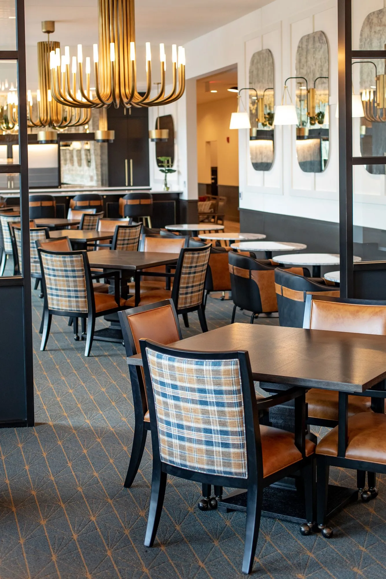 Senior living dining room with plaid and leather chairs at dark tables under brass candle chandeliers, arched mirrors beyond