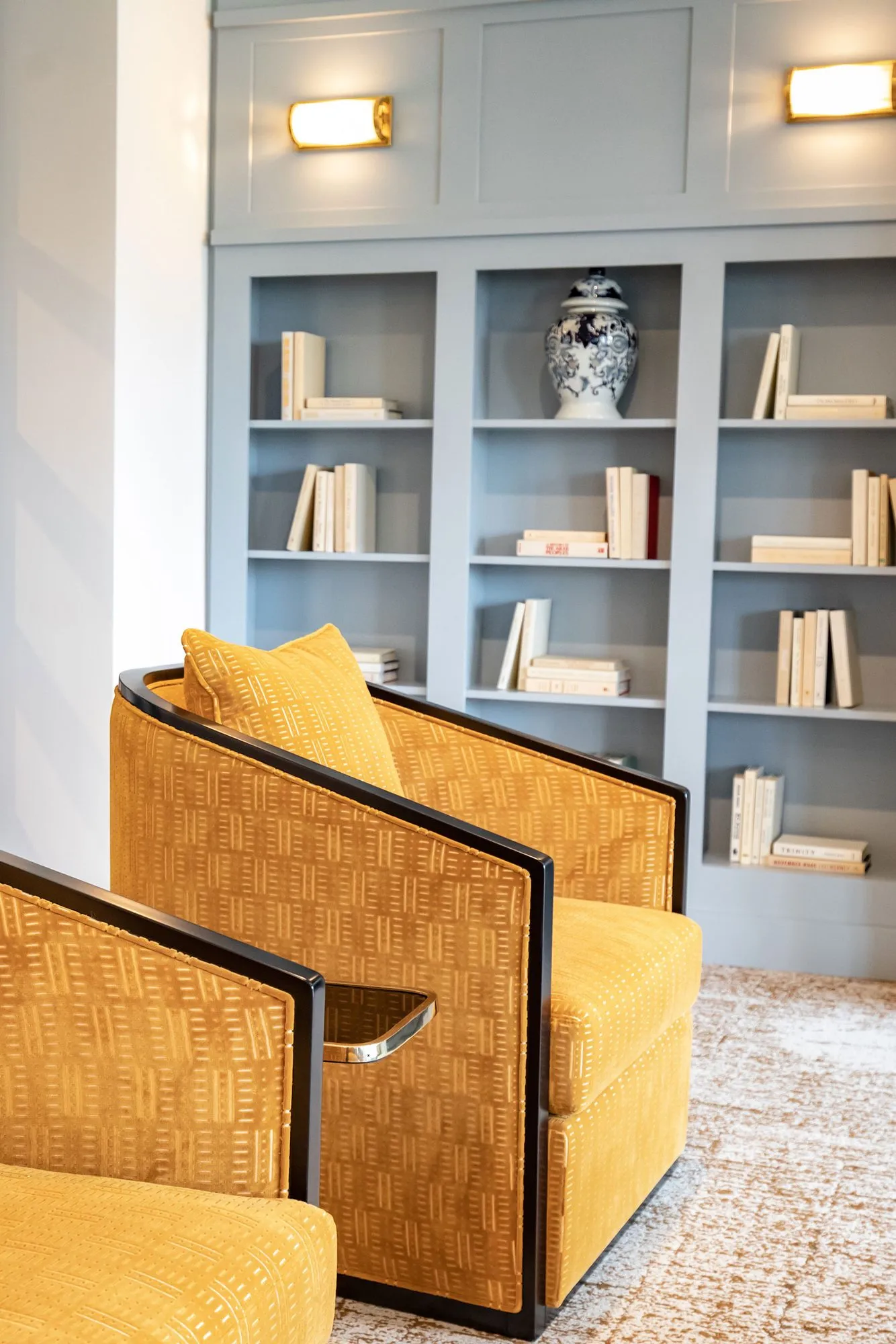 Senior living interior detail: mustard patterned swivel chairs with black frames before a pale blue bookcase and ginger jar