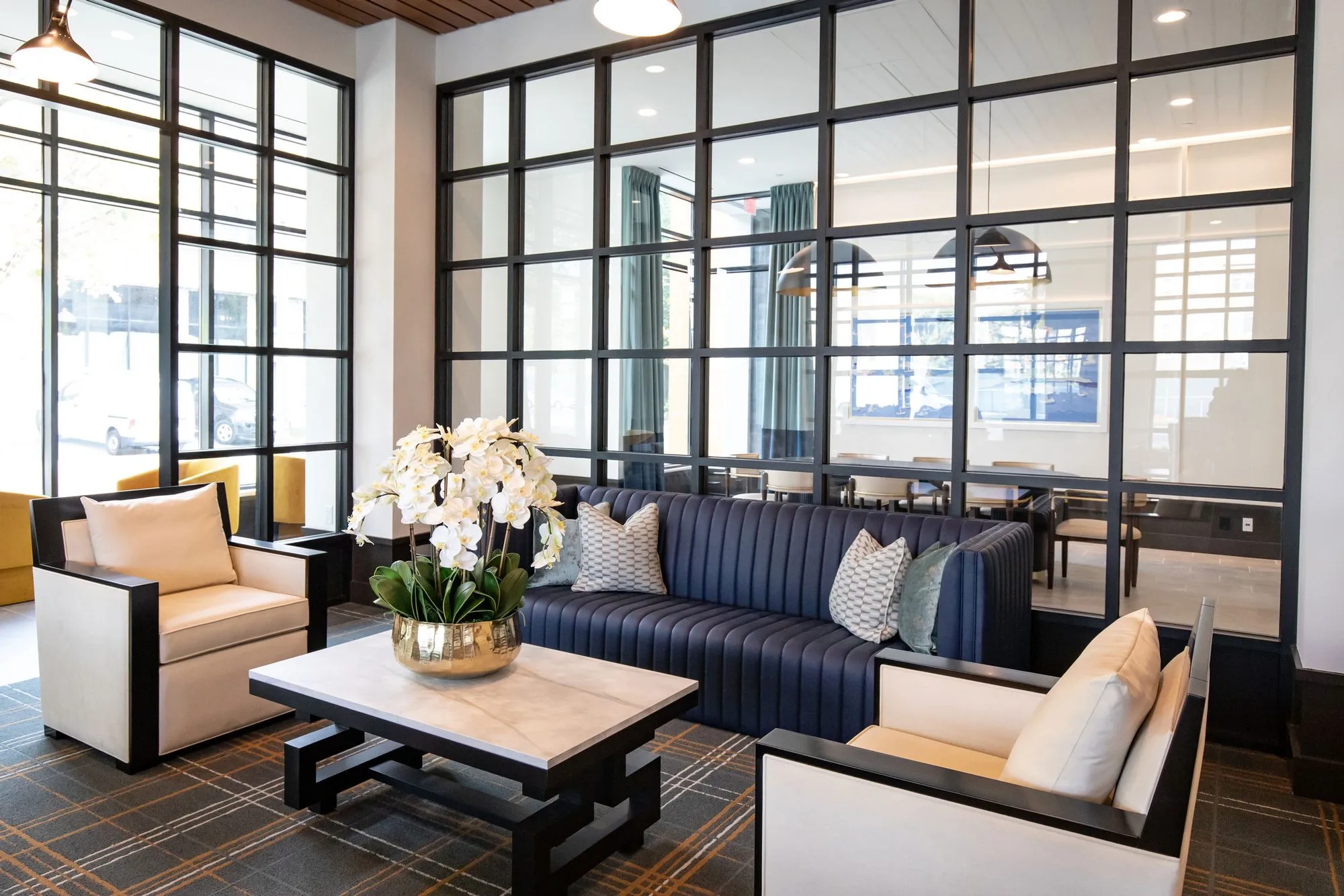 Senior living lounge with a navy channel tufted sofa and paired cream armchairs around a marble table, steel glazing behind