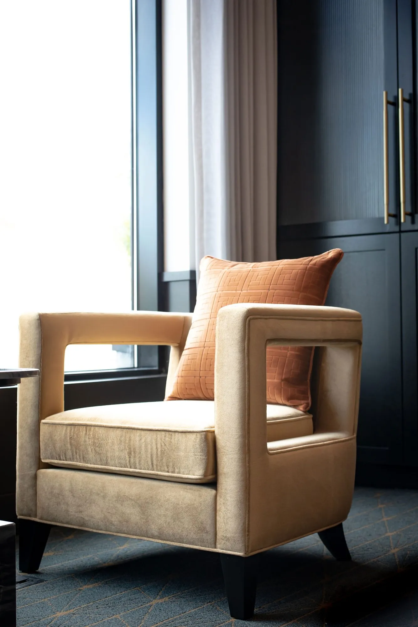 Senior living interior detail: camel velvet armchair with open arms and an orange plaid pillow by a window and navy cabinetry