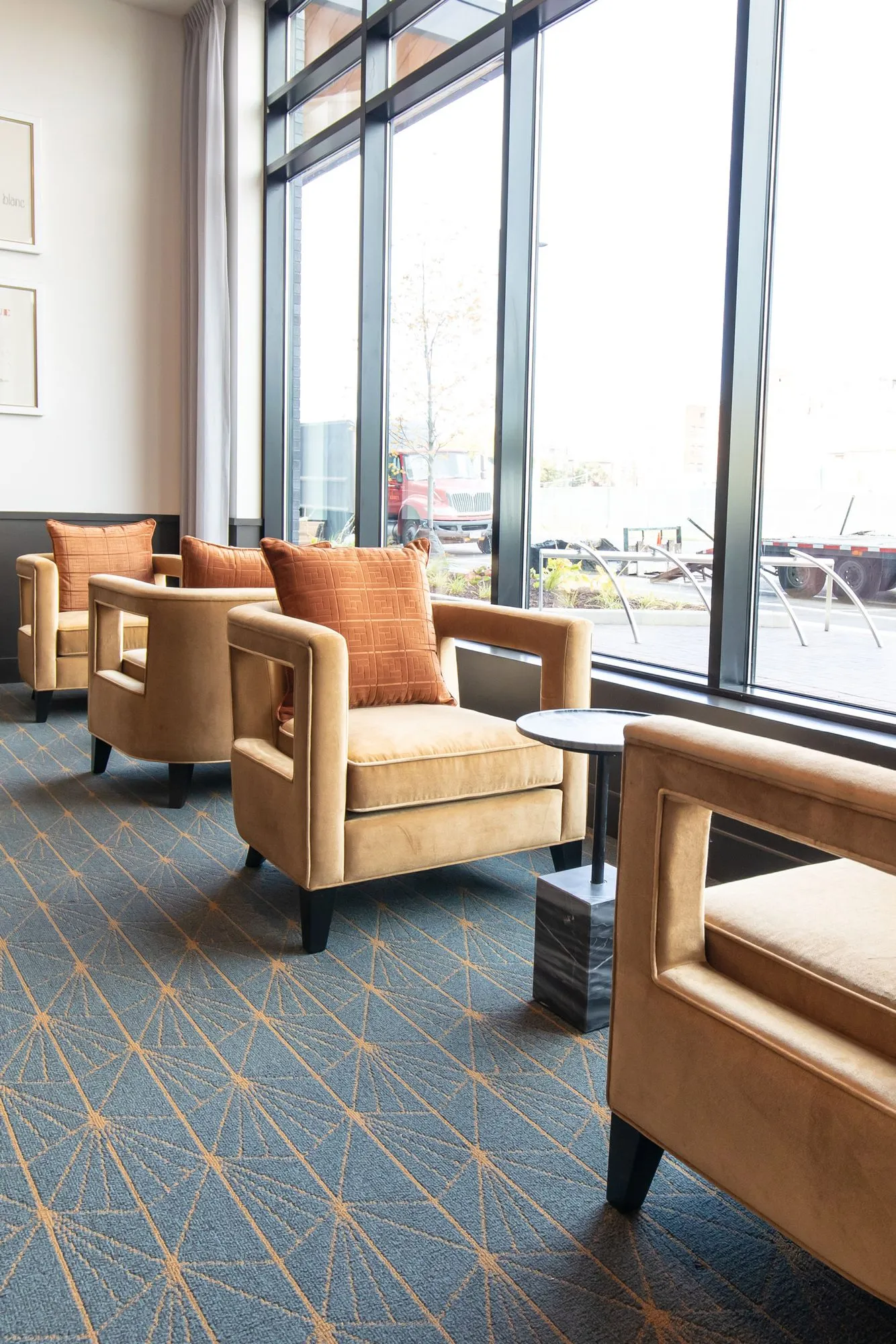 Senior living sitting area with a row of camel velvet chairs with orange pillows along tall windows on starburst carpet