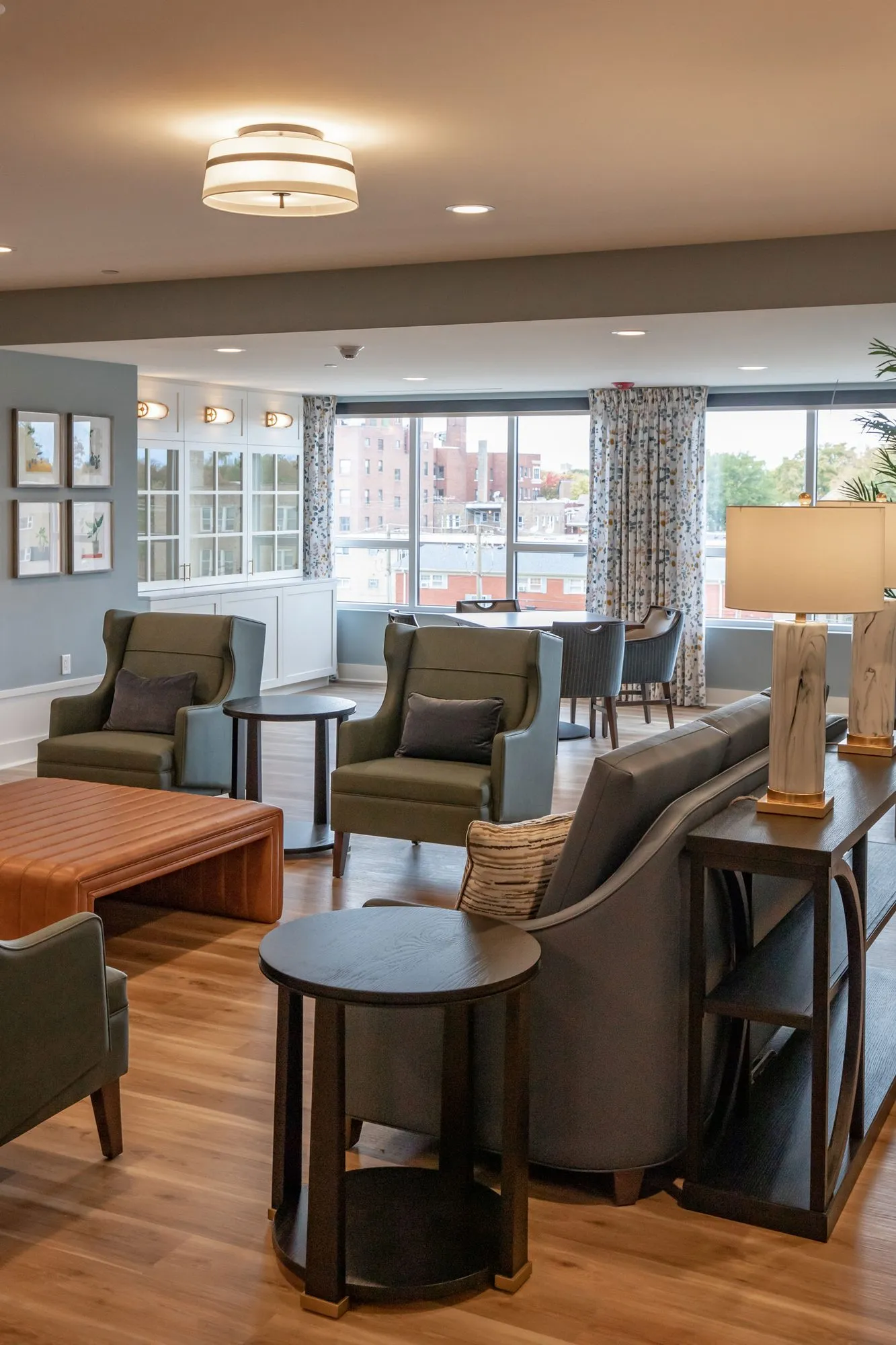 Senior living lounge with sage wingback chairs, an orange leather ottoman and a grey sofa backed by a lamp console