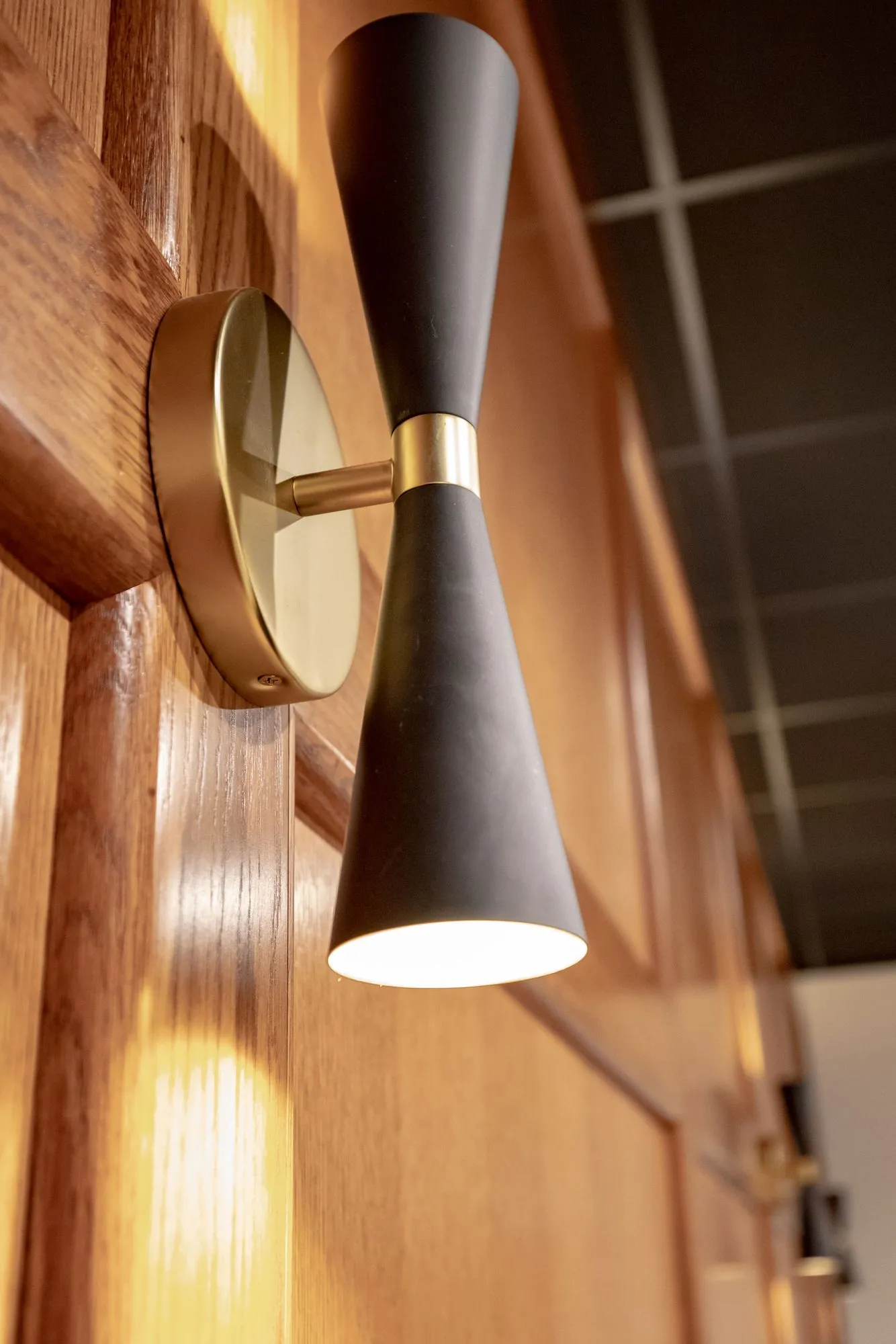 Senior living interior detail: black and brass hourglass wall sconce on wood panelling, the dark ceiling grid beyond