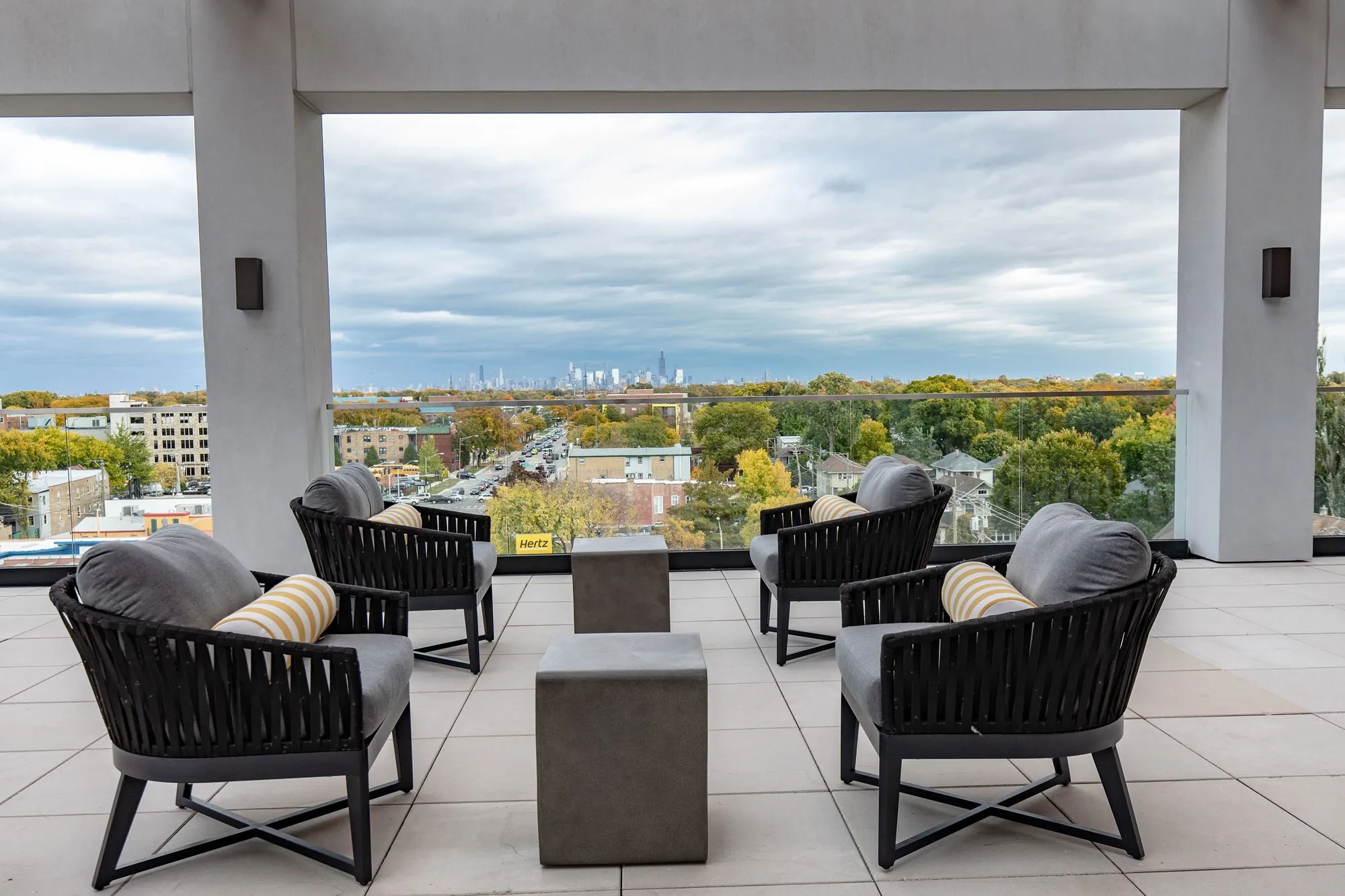 Senior living rooftop terrace with black rope lounge chairs and concrete cube tables between columns, city skyline beyond