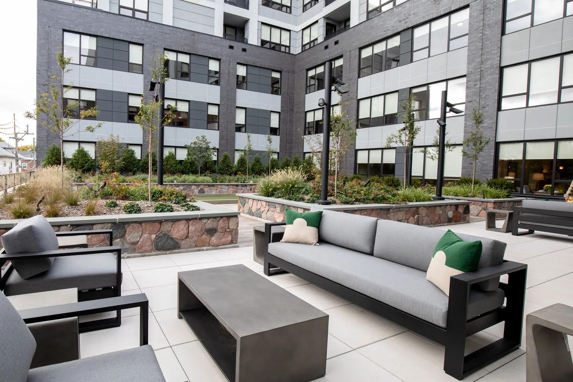 Senior living outdoor seating: grey sofa with green pillows and a concrete coffee table, lamp posts and planted beds beyond