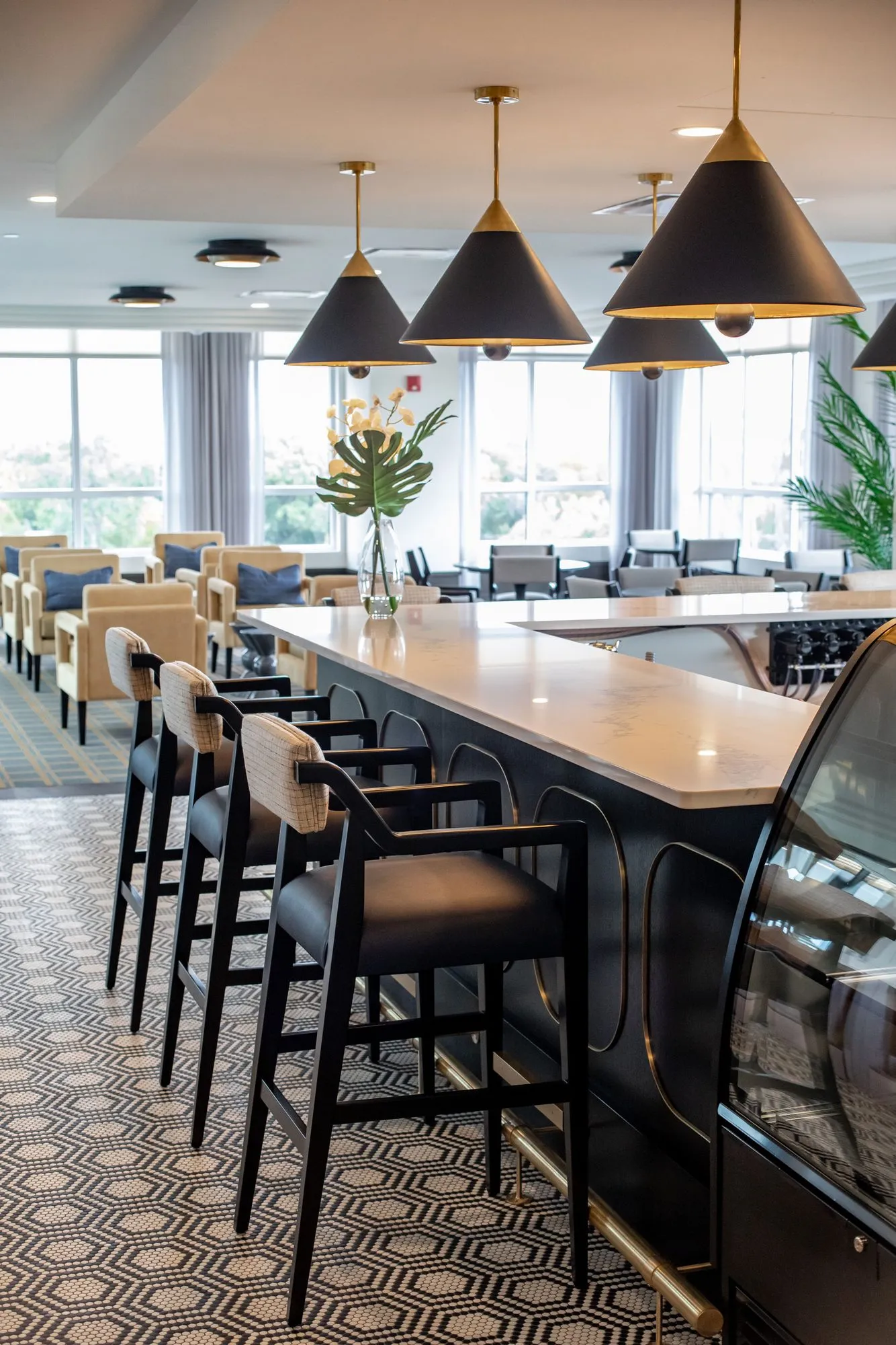 Senior living bistro bar with cane back stools under black cone pendants, camel lounge chairs by the windows beyond
