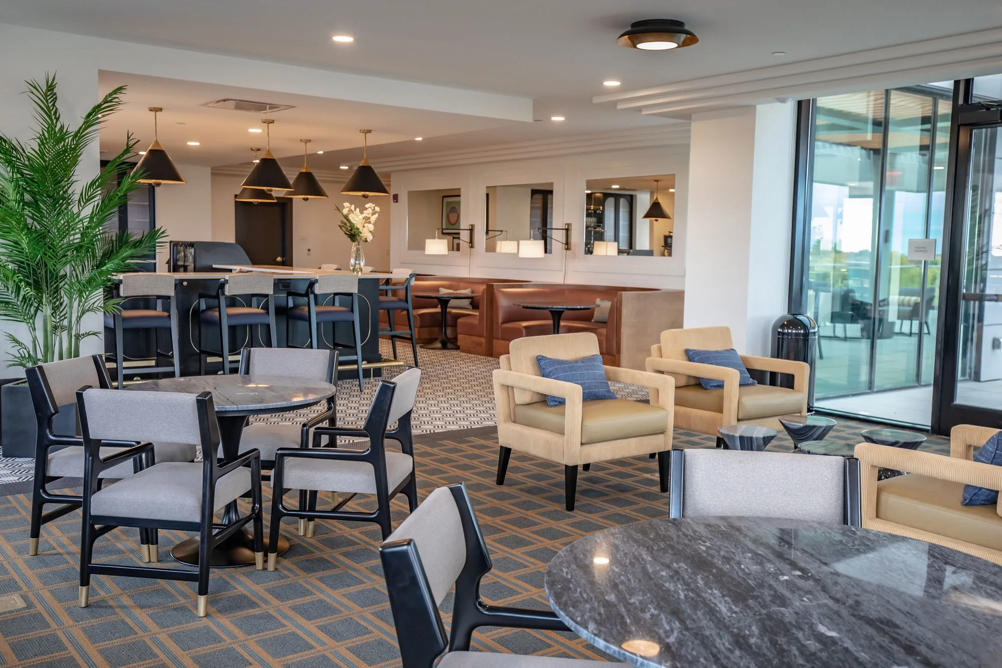 Senior living bistro seating of camel armchairs and dark chairs at a marble table, bar and banquettes beyond