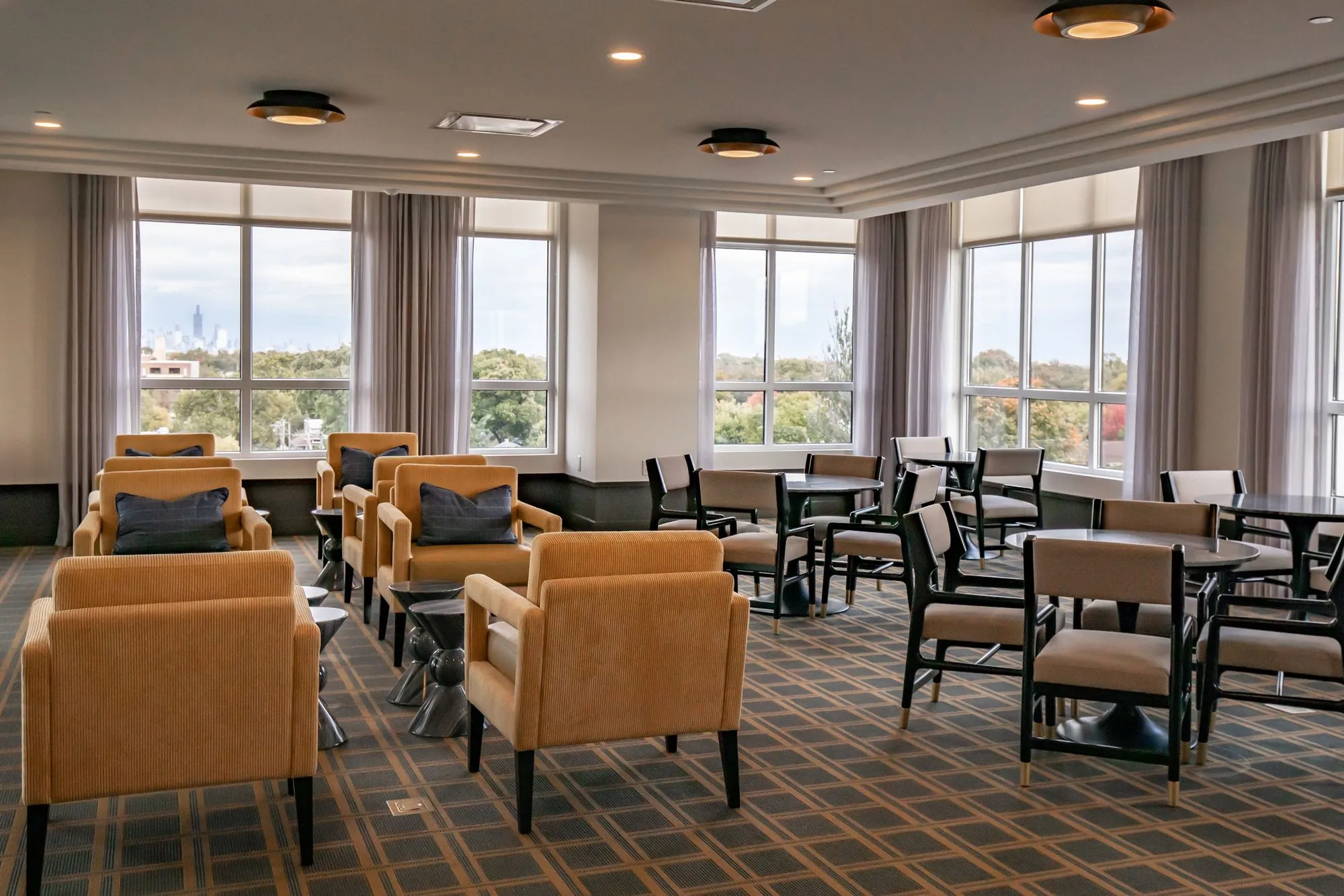 Senior living lounge with camel armchairs and dining tables on a geometric carpet below tall curtained windows