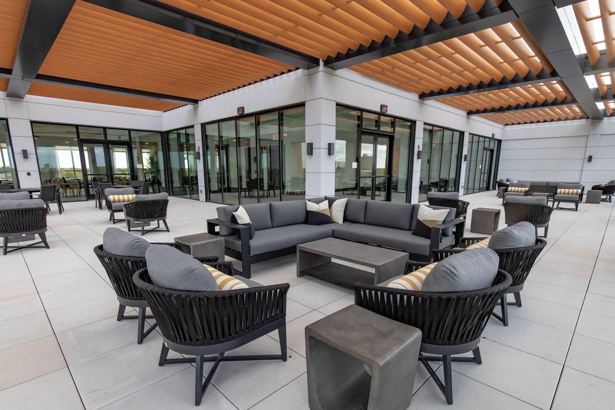 Senior living roof terrace under slatted timber canopies, grey sectional and rope chairs on pale pavers
