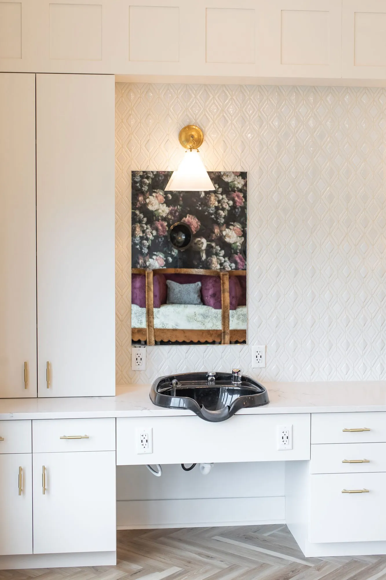 Senior living salon detail: black shampoo basin on a white vanity below a brass sconce and mirror reflecting floral wallpaper