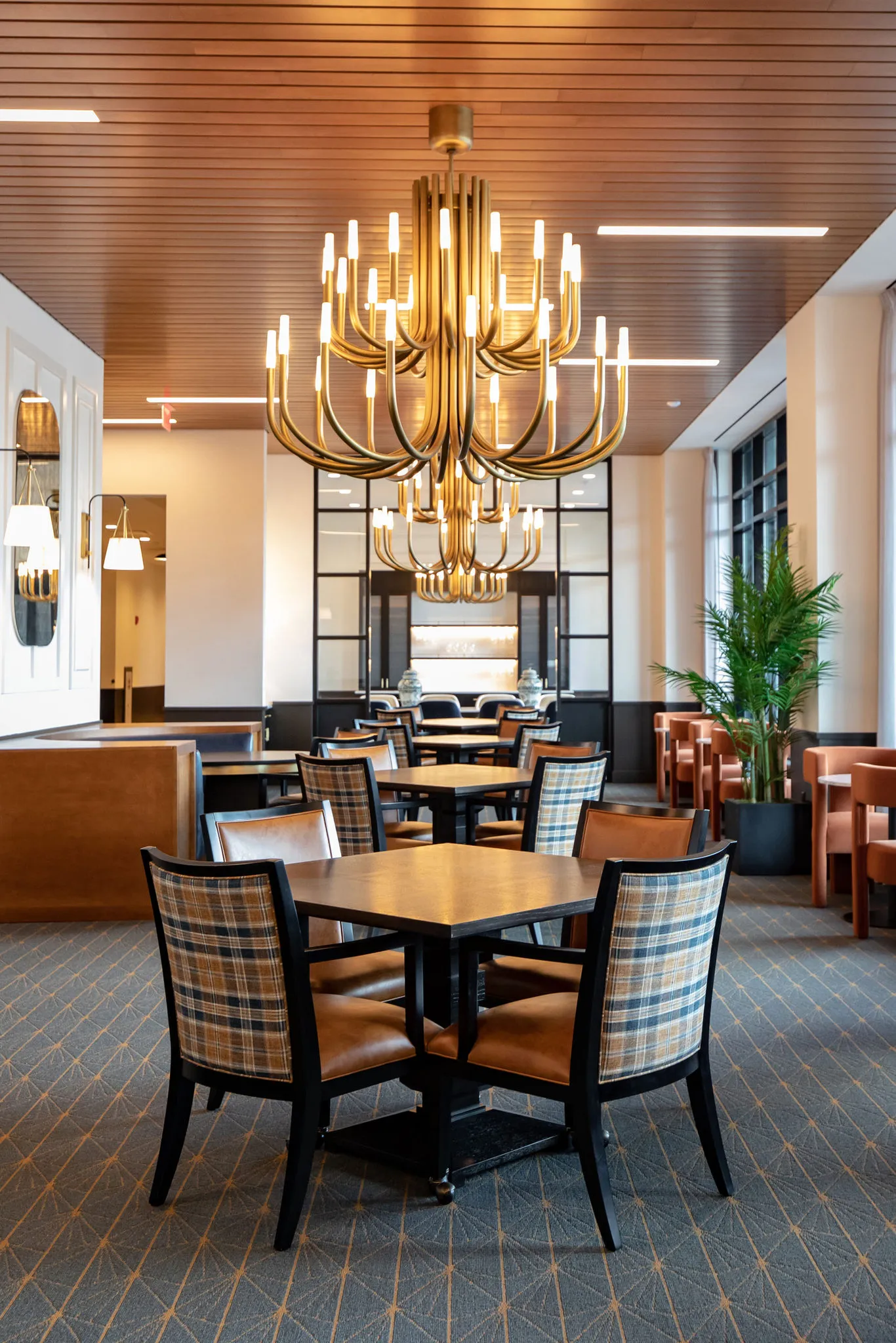 Senior living dining room with brass chandeliers below a wood slat ceiling above plaid and leather chairs