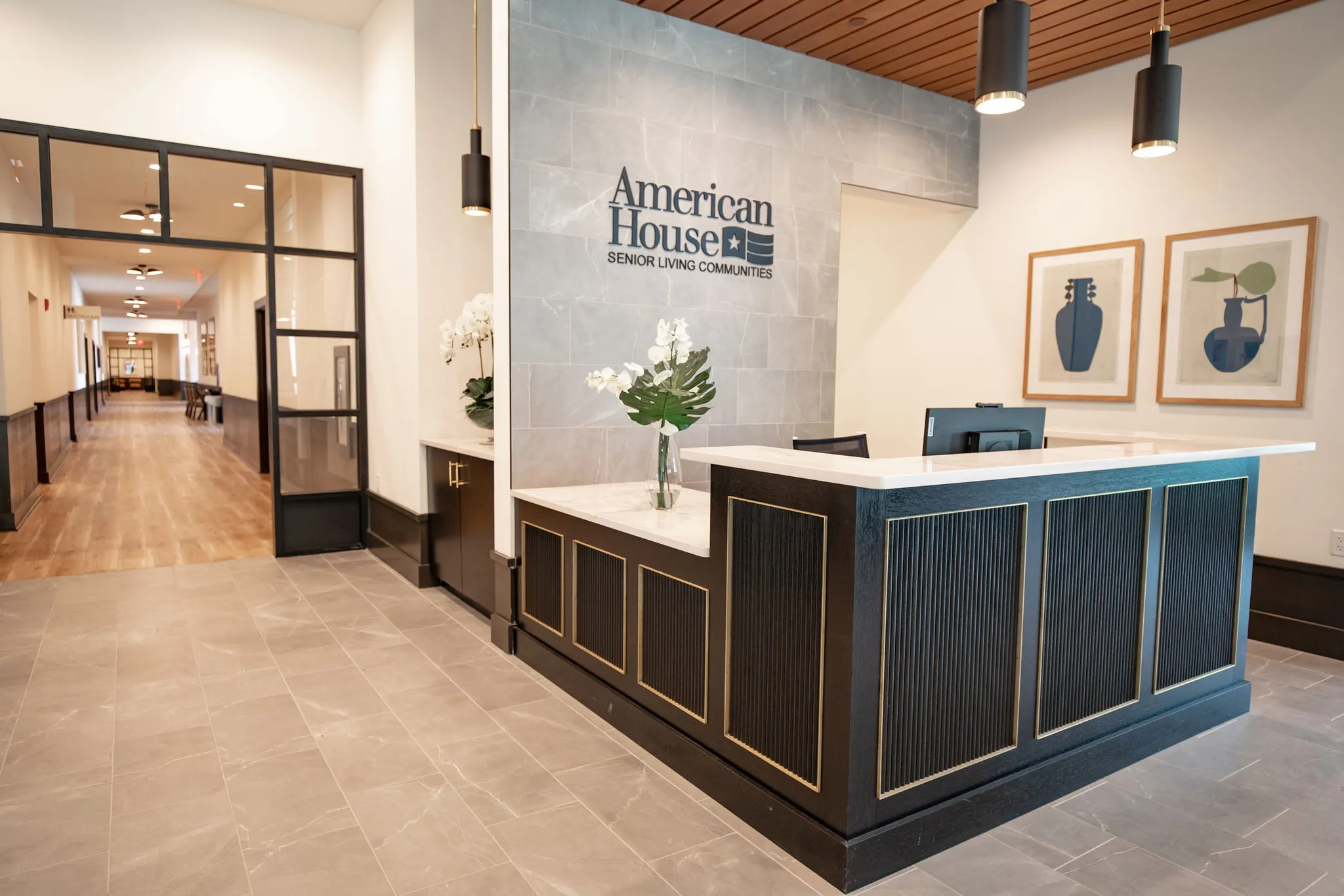 Reception lobby with a navy and brass front desk, a grey stone feature wall, white orchids and a corridor of black steel glazing beyond