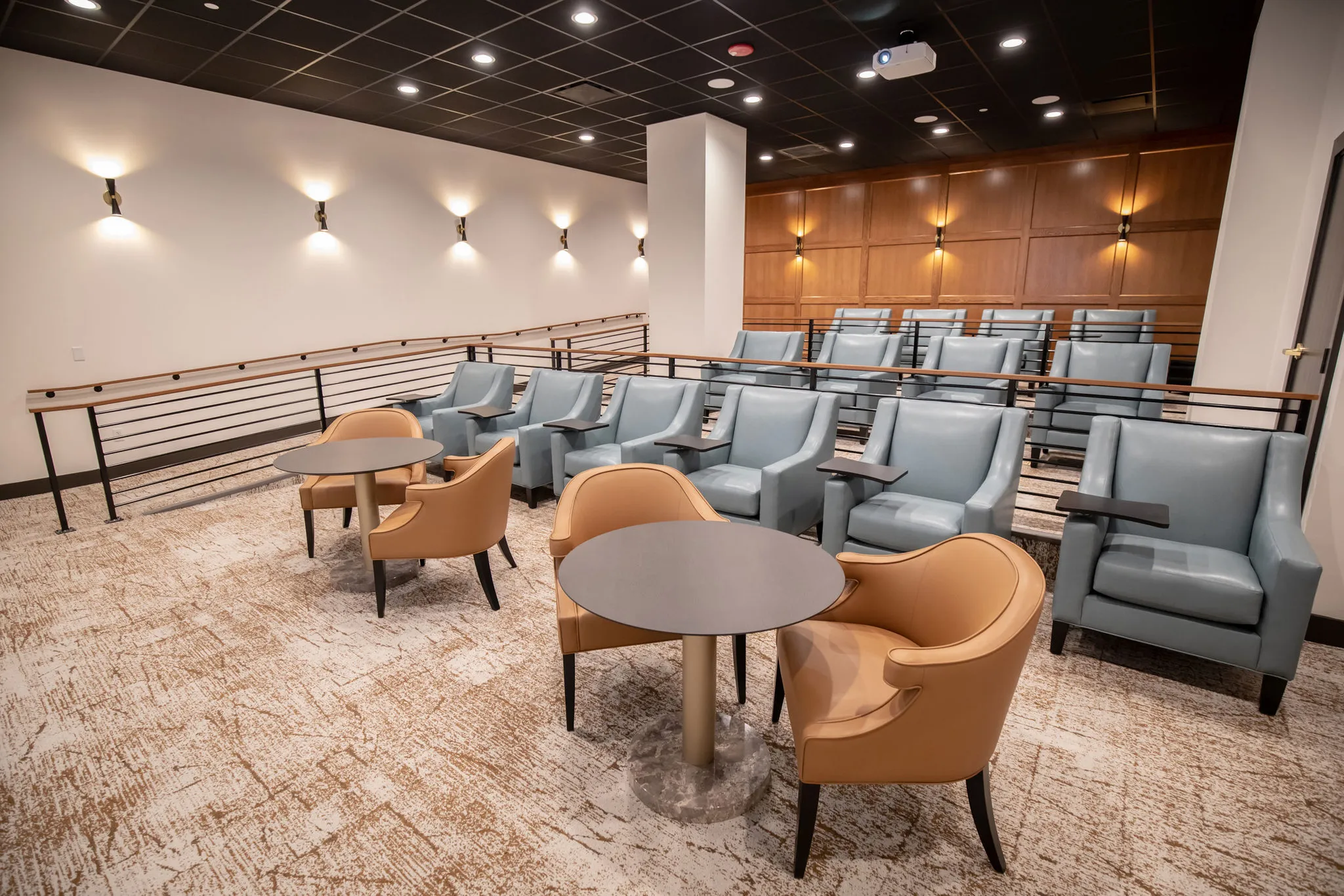 Senior living theater with tiered rows of blue-grey recliners behind camel chairs at cafe tables, a walnut panelled back wall