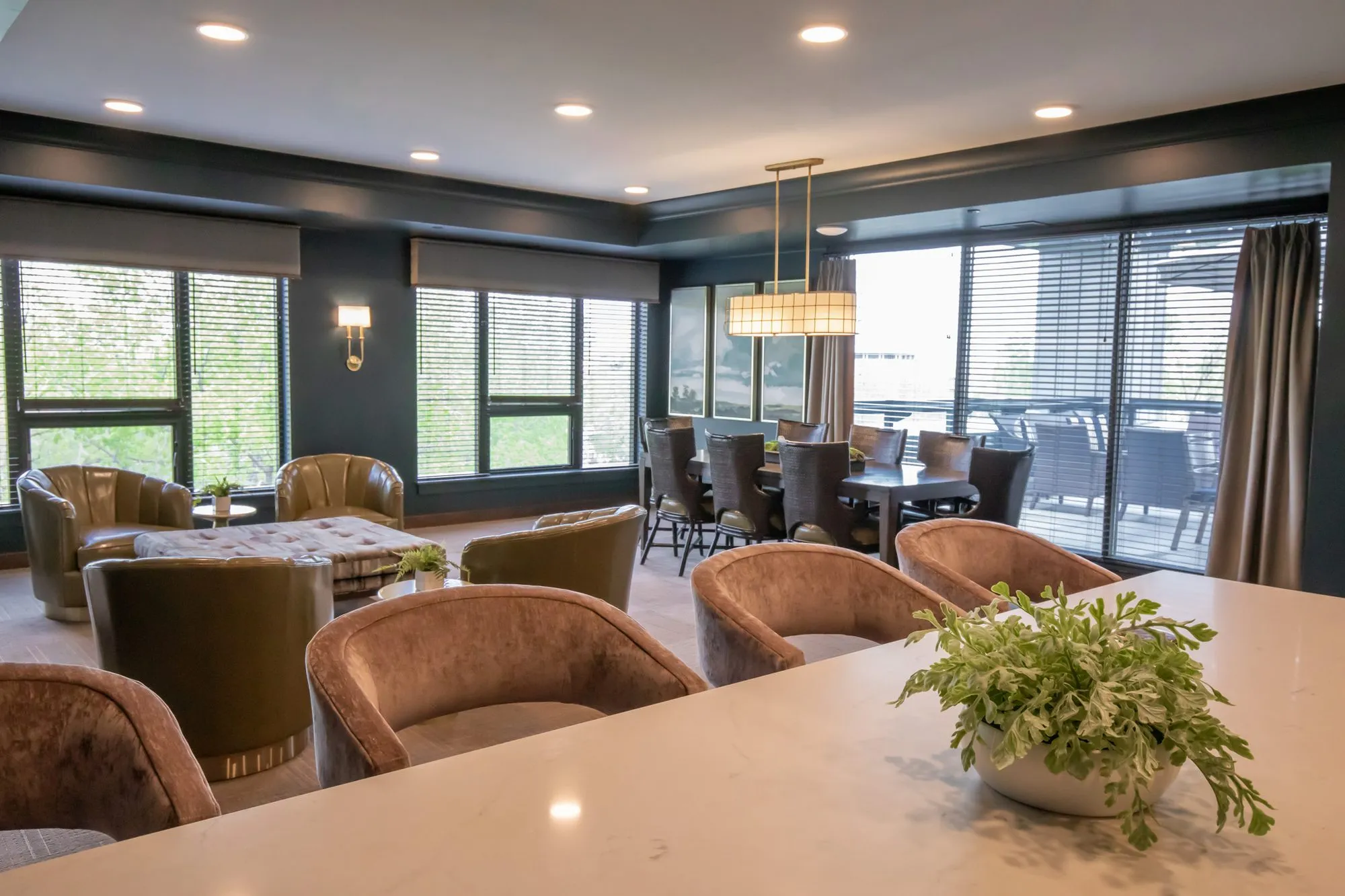 Active adult community lounge of blush velvet tub chairs and olive armchairs beside a dining area under a linear pendant